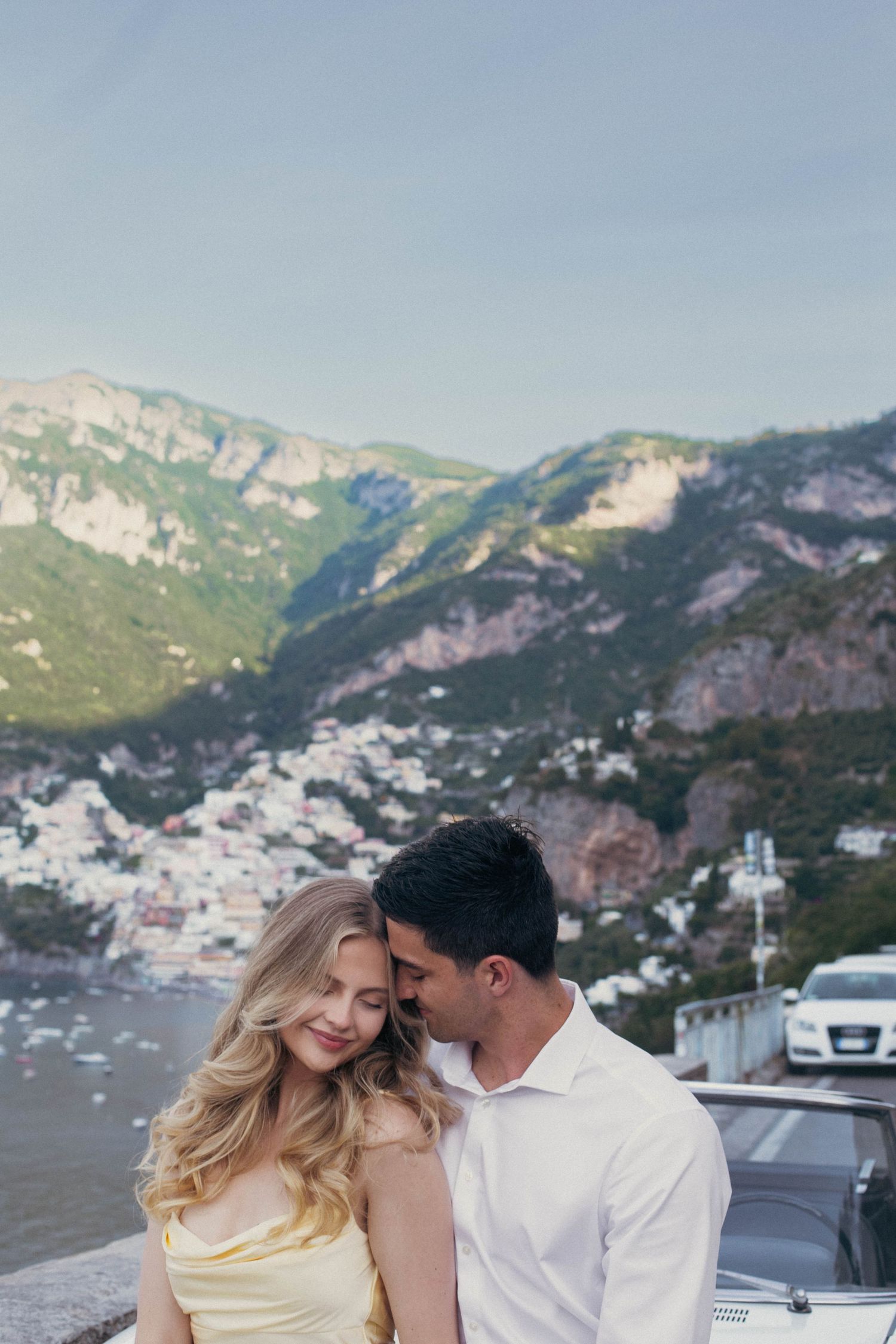 Romantic couple embracing on Amalfi Coast overlook with Positano village and green mountains behind them.