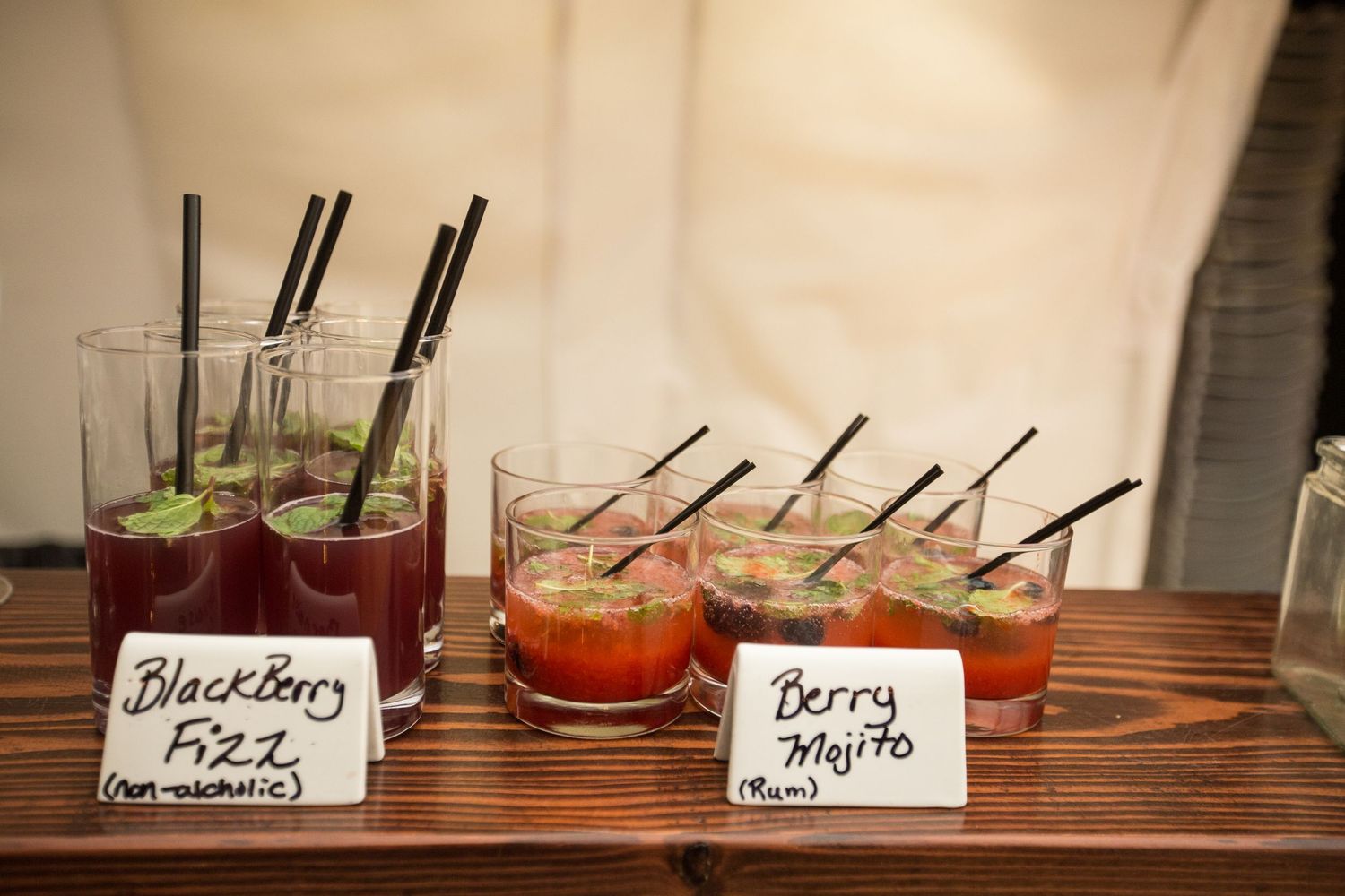 Blackberry Fizz and Berry Mojito cocktails displayed on a wooden bar with handwritten signs at an event.