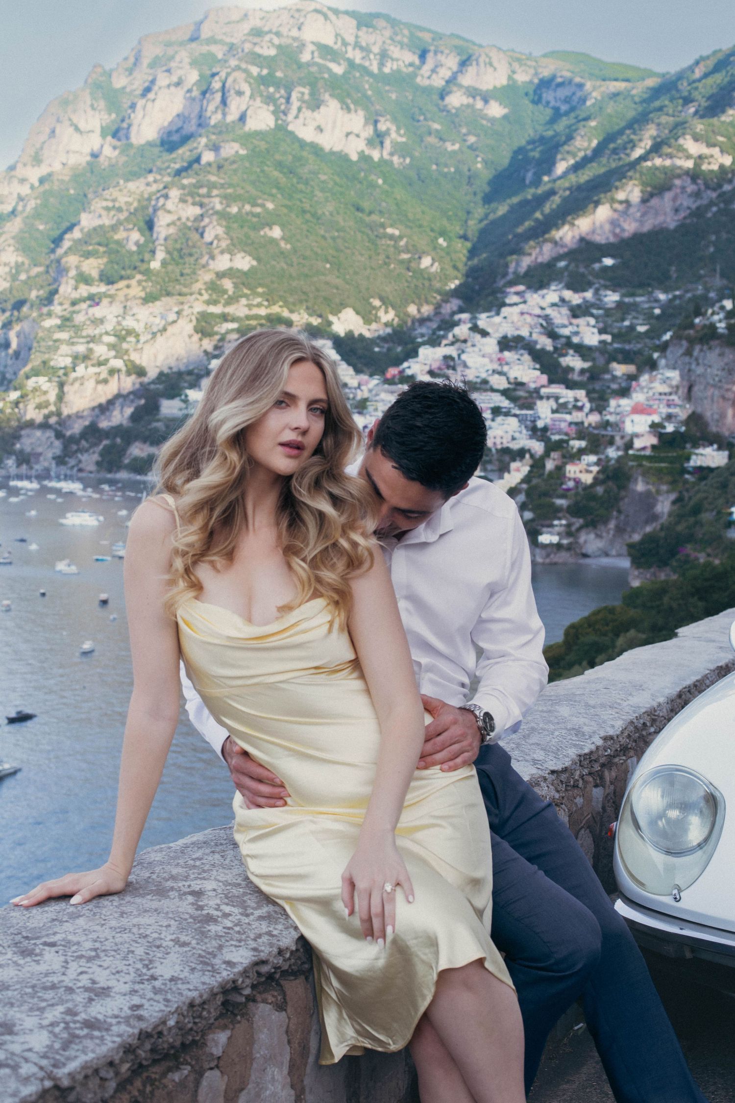Glamorous blonde woman in yellow satin dress with man embracing her against Amalfi Coast backdrop.