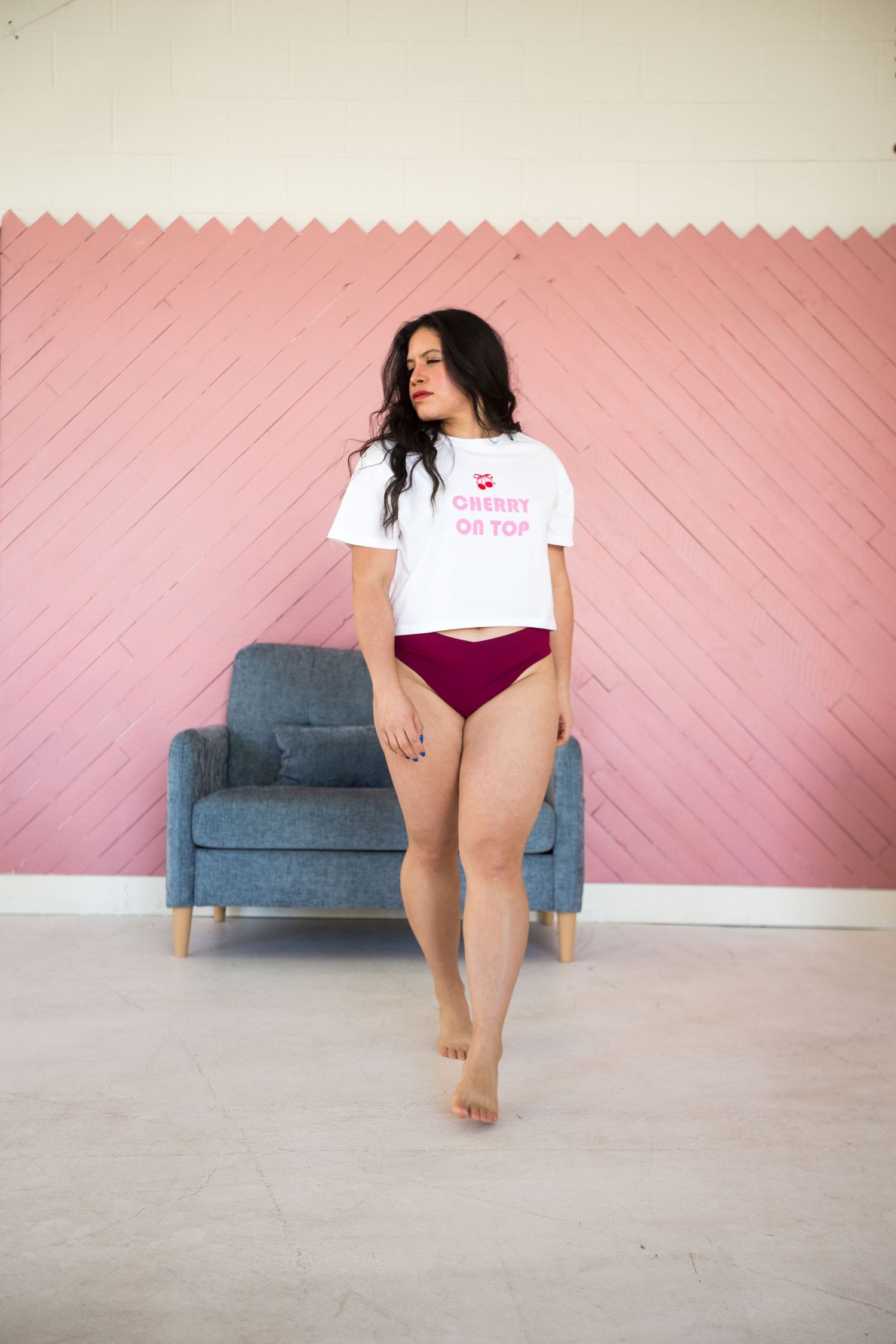 Person in white t-shirt and burgundy underwear poses near blue couch against pink wall.