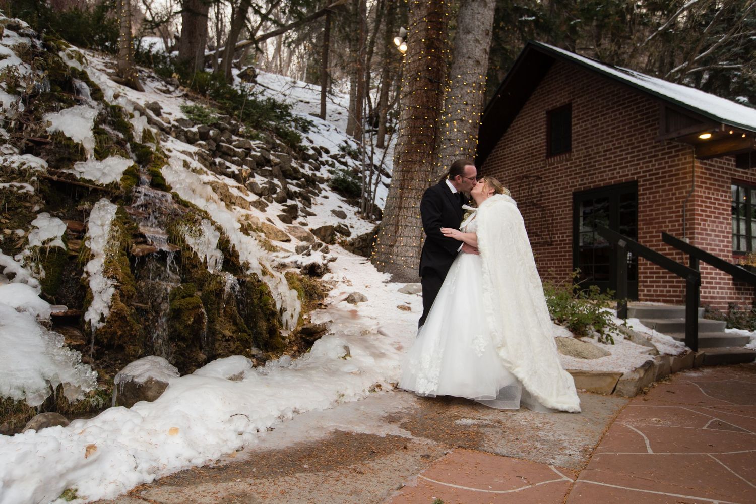 Wedding couple poses against brick building backdrop with snow-covered ground and trees.