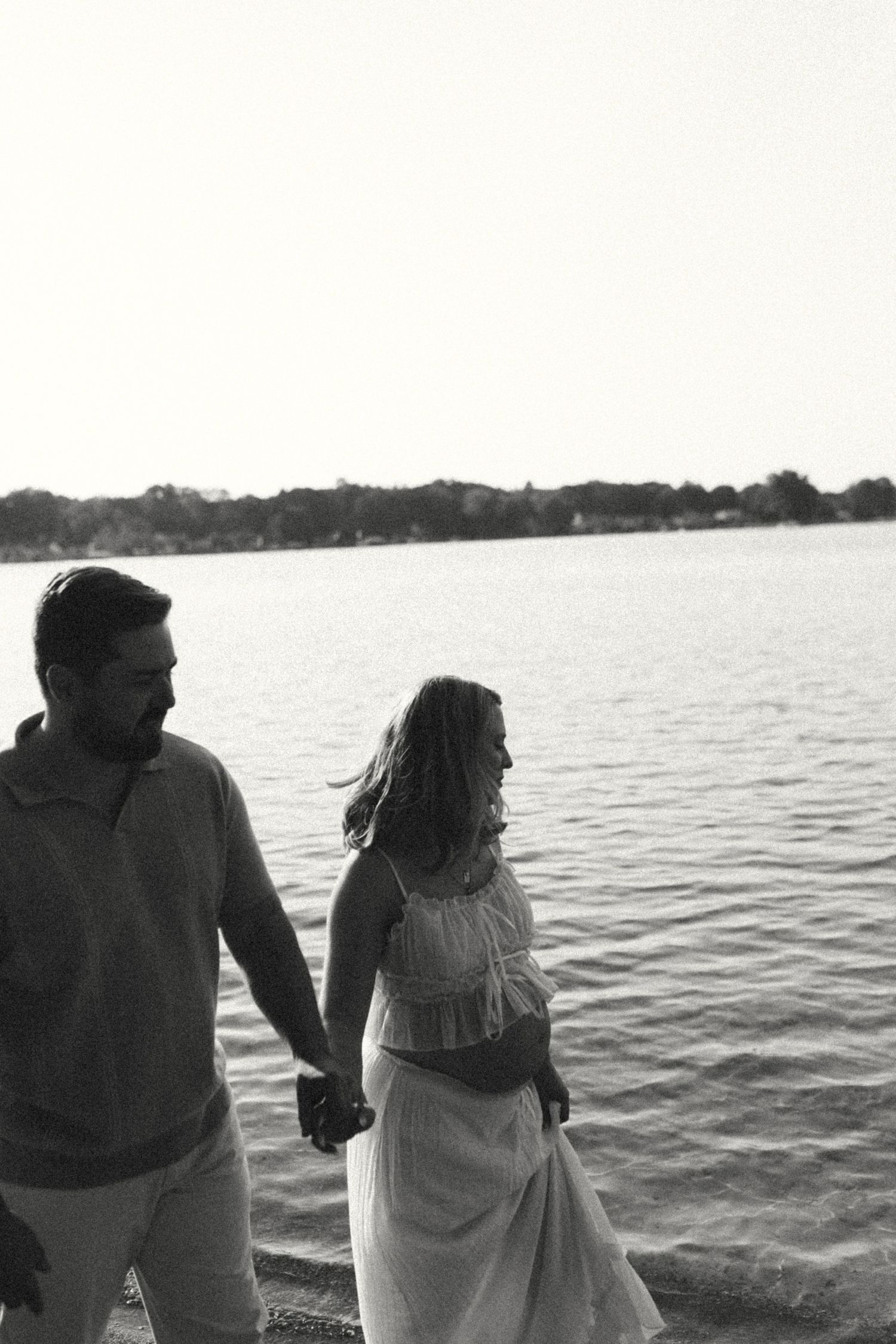 A couple holding hands walking near a lake at sunset in a black and white photo.