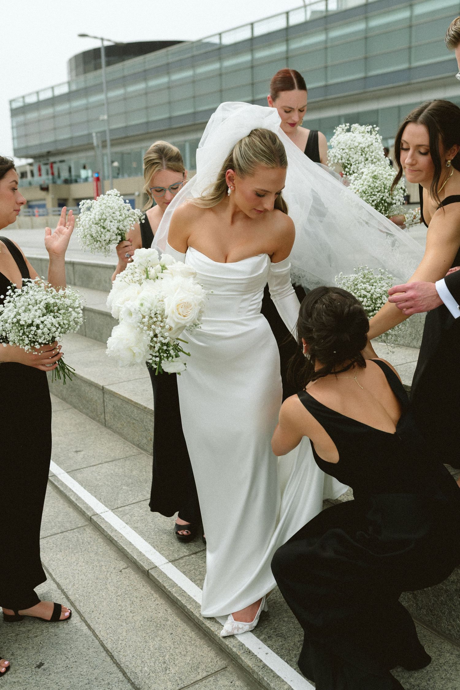 Bridesmaids in black dresses adjust bride's white gown and veil on outdoor urban staircase.