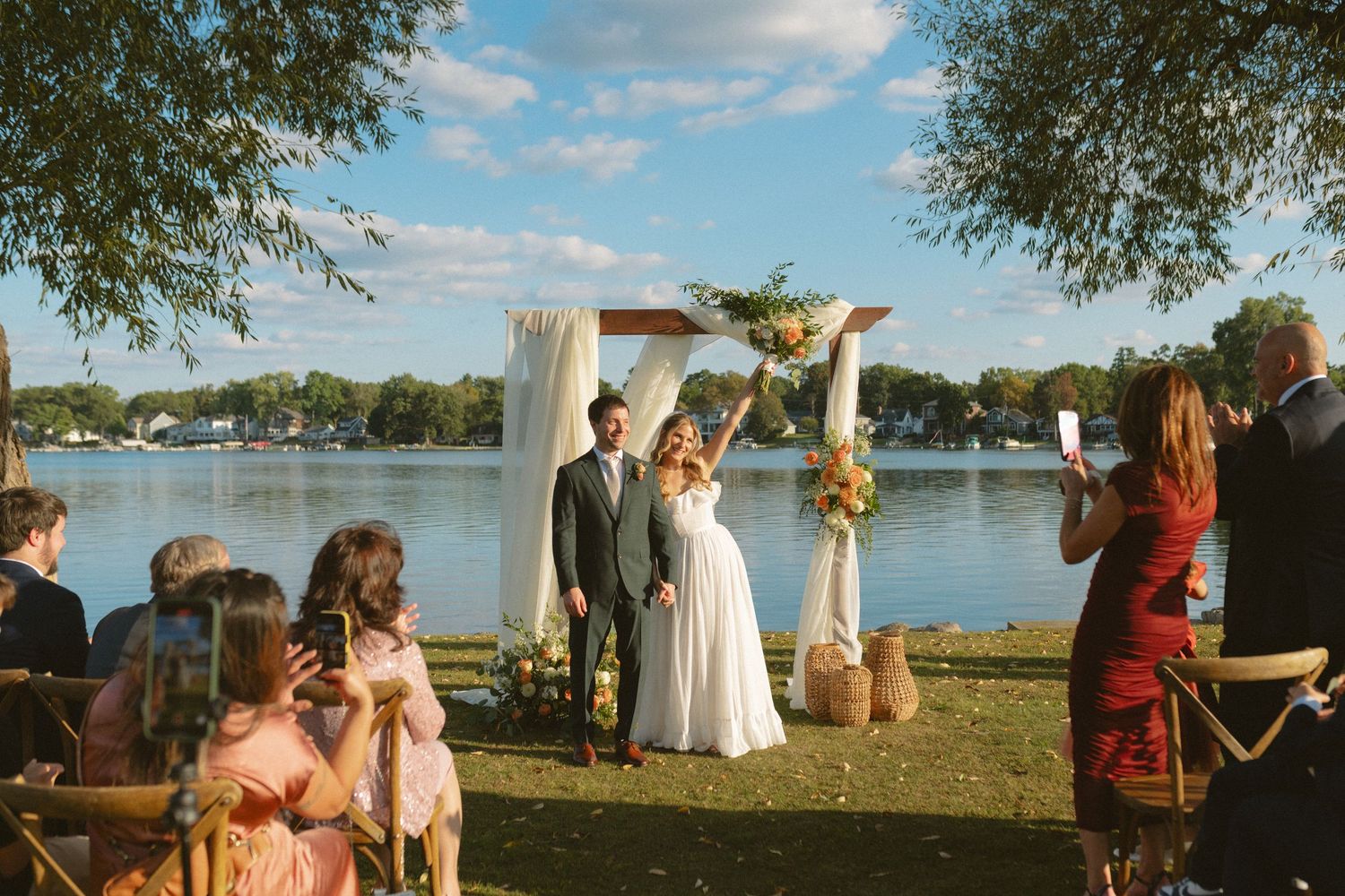 Bride and groom celebrate at outdoor lakeside wedding ceremony under rustic floral arch on sunny day.