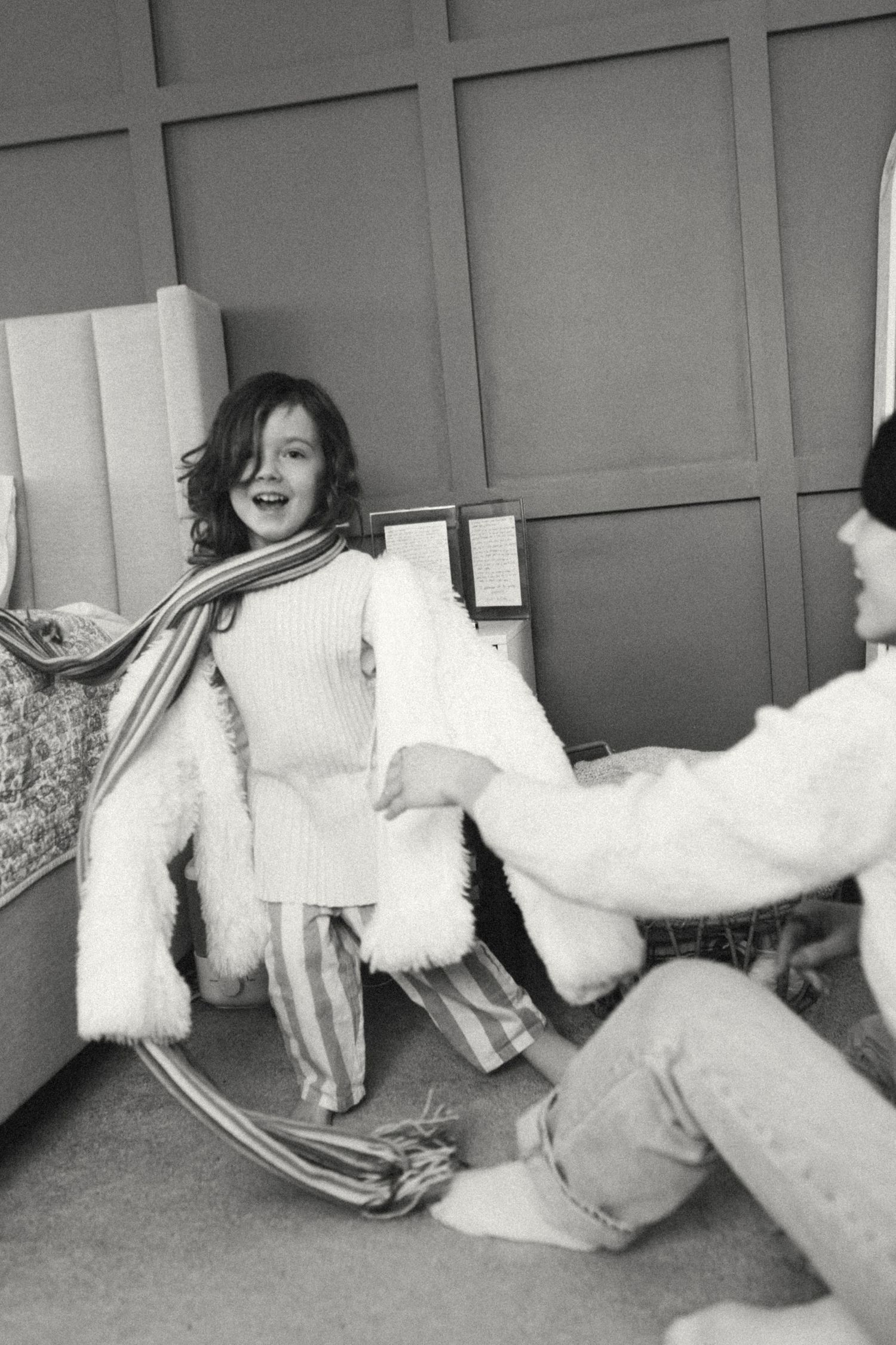 Black and white photo of smiling child in fluffy coat and striped scarf playing with adult indoors.