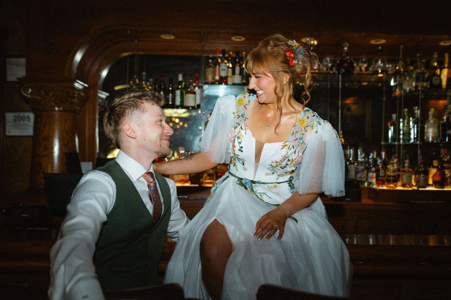 Bride in floral dress laughing with groom at a bar during wedding reception celebration.