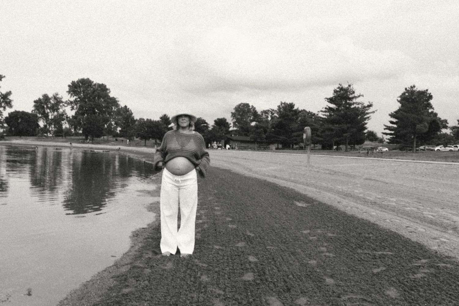 Black and white photo of pregnant woman standing by lakeside in white pants and straw hat.