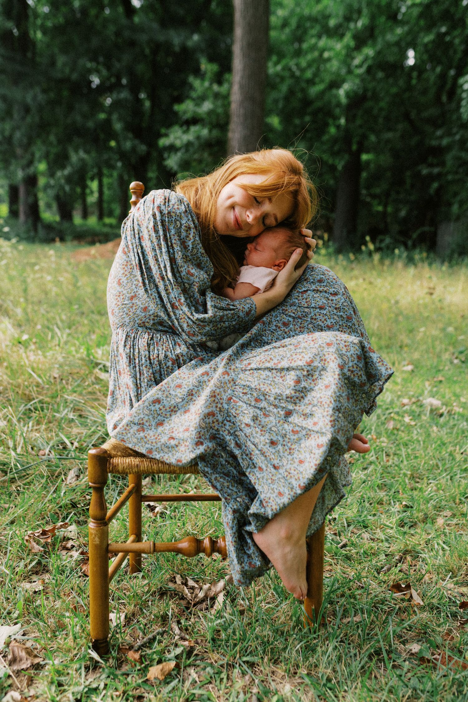 Red-haired mother sitting on wooden chair outdoors, tenderly holding newborn baby wrapped in floral shawl in green park.