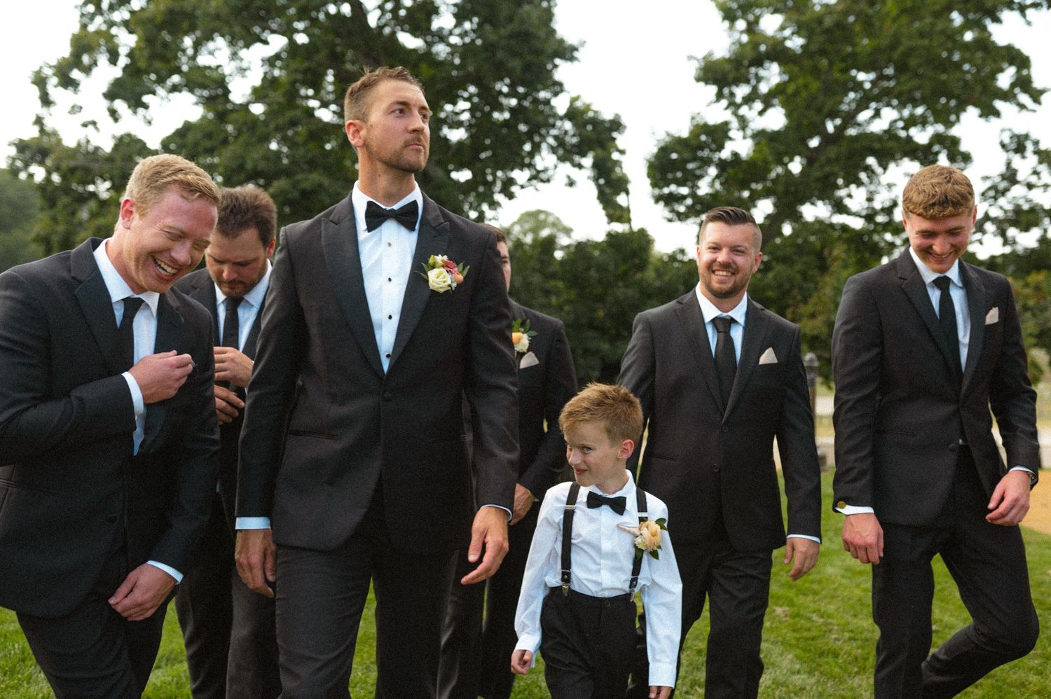 Groom in black tuxedo laughing with groomsmen and ring bearer in suspenders outdoors.