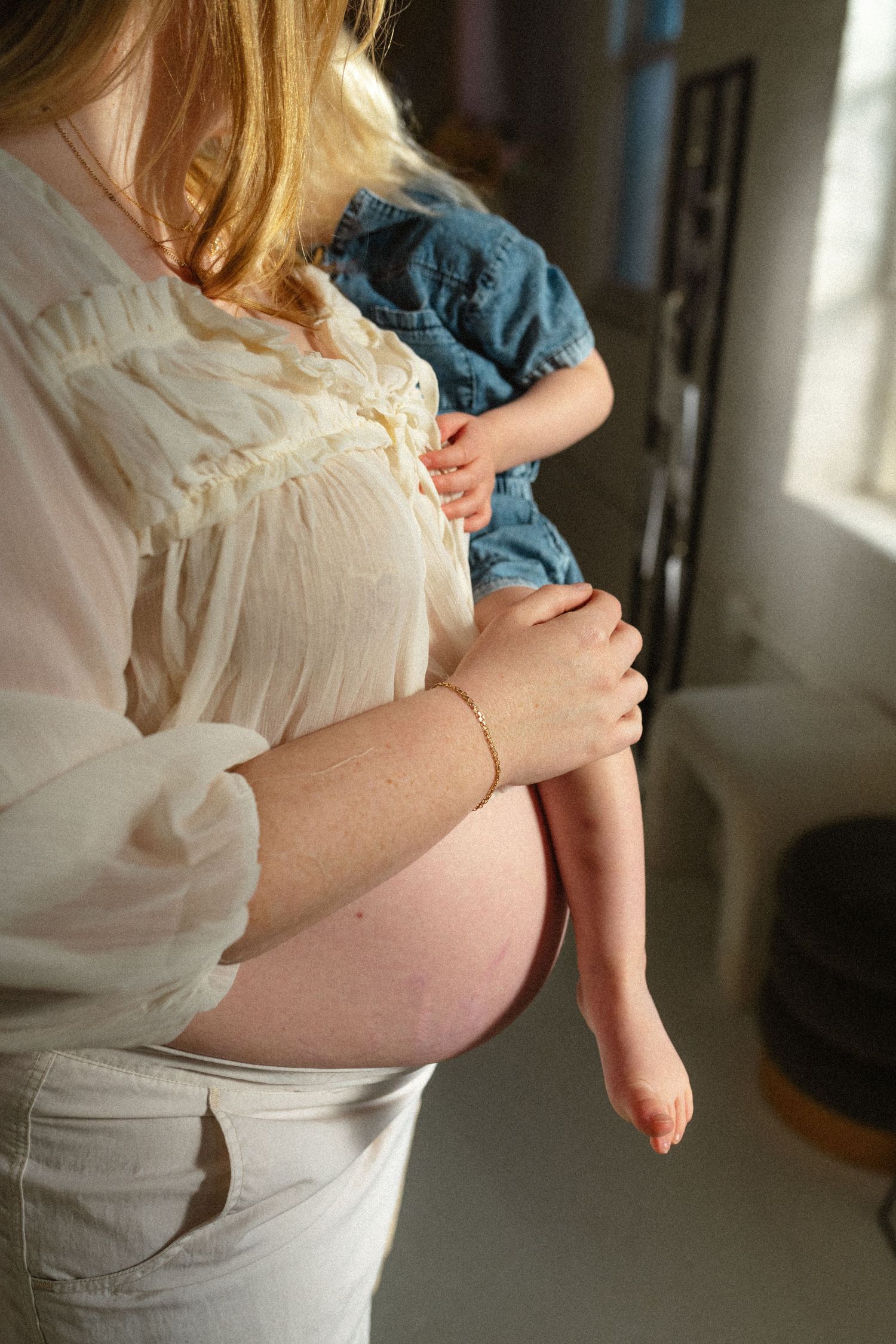 Pregnant woman in white blouse holding toddler child against her baby bump indoors.