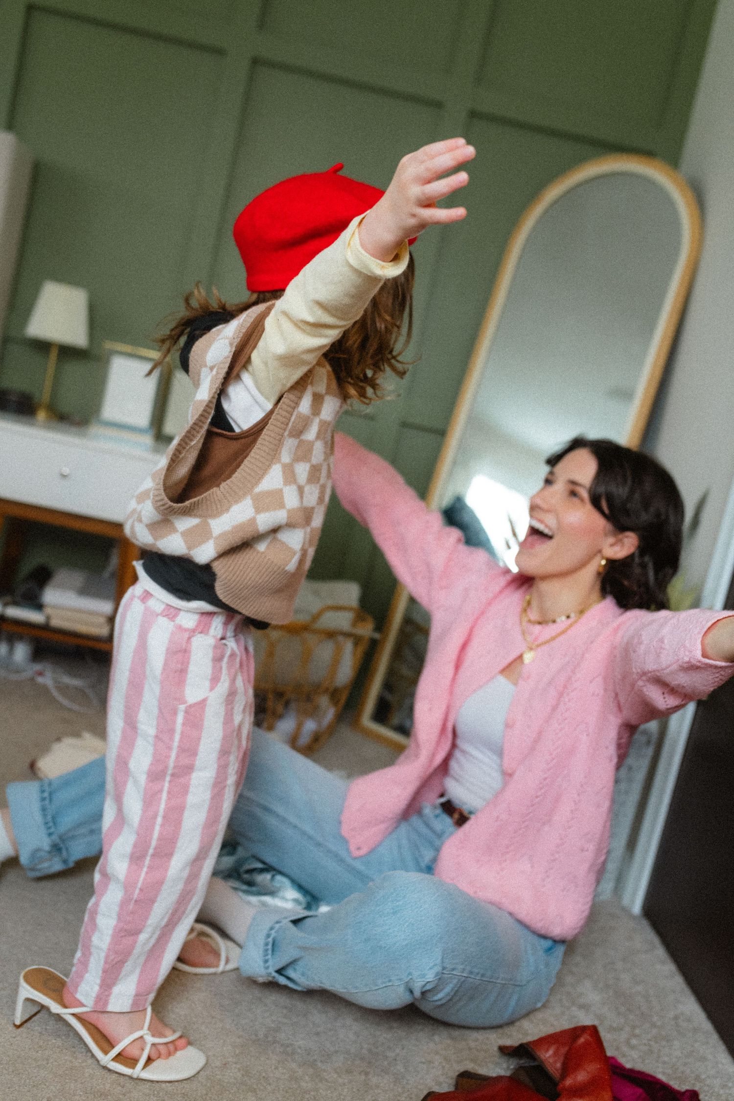 Excited mother in pink cardigan playing with daughter in red beret and striped pants in bedroom.