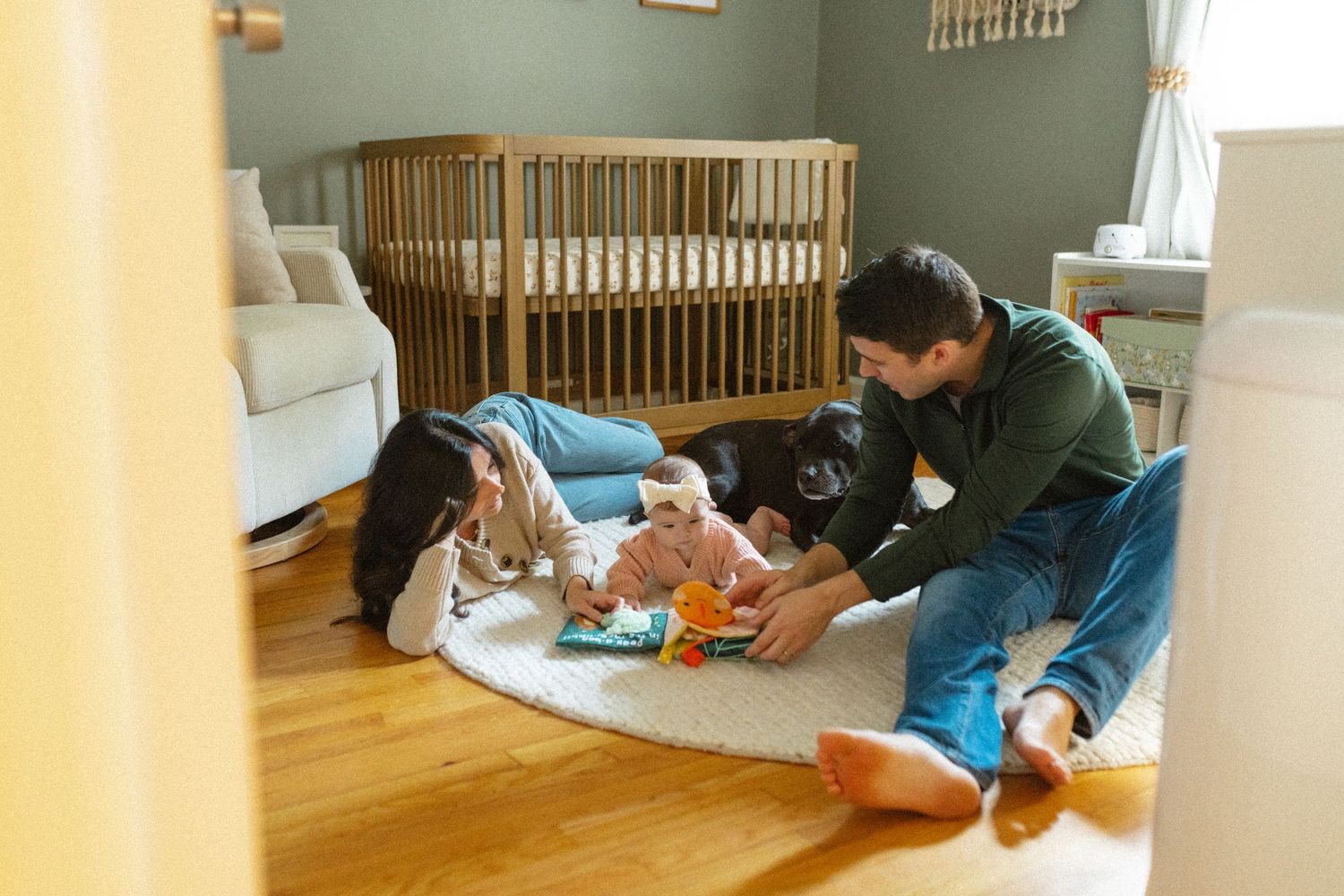 Parents and baby play on nursery floor with a black dog beside a wooden crib.