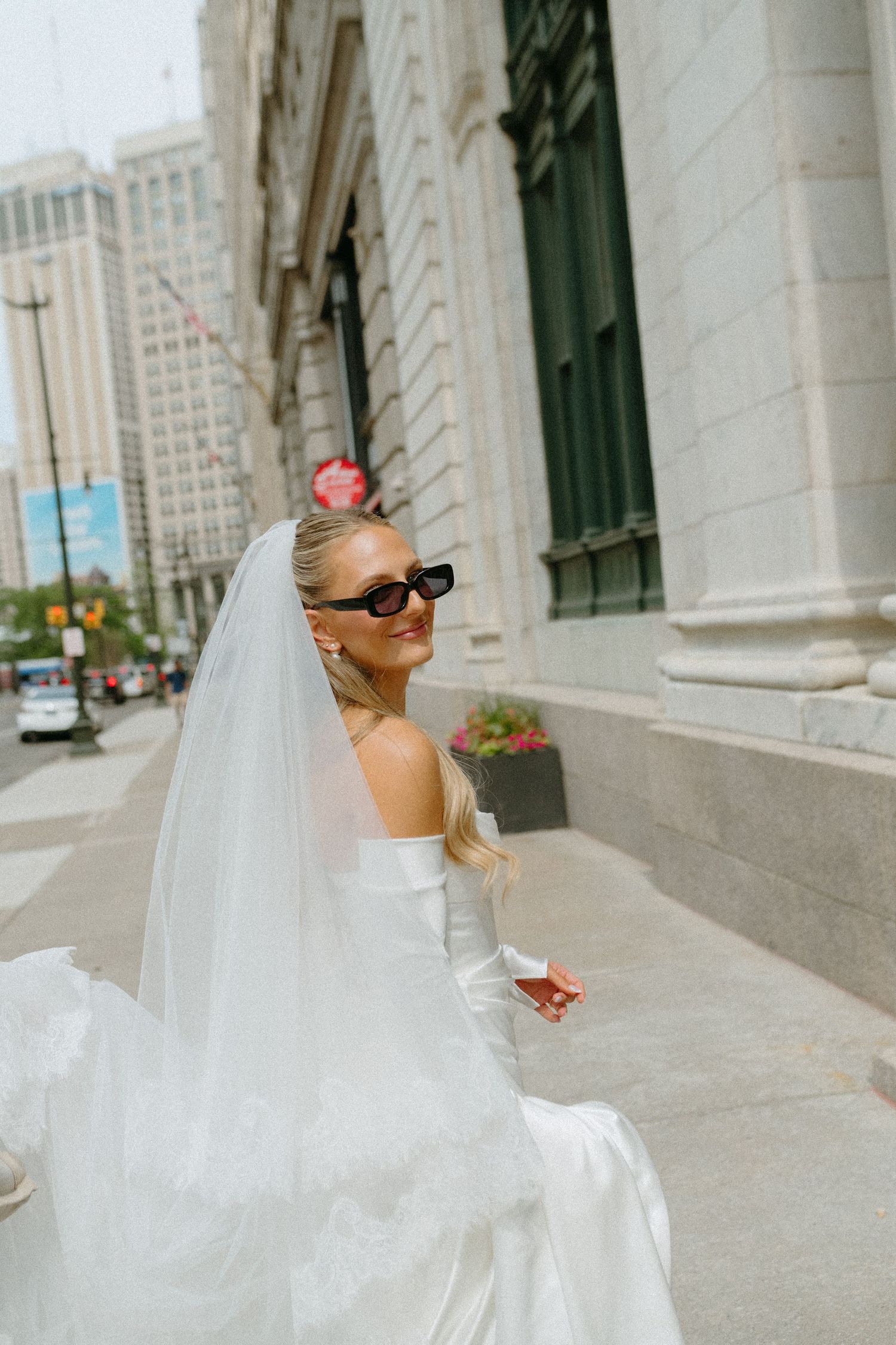 Bride in white strapless gown and veil wearing sunglasses poses on urban city sidewalk.