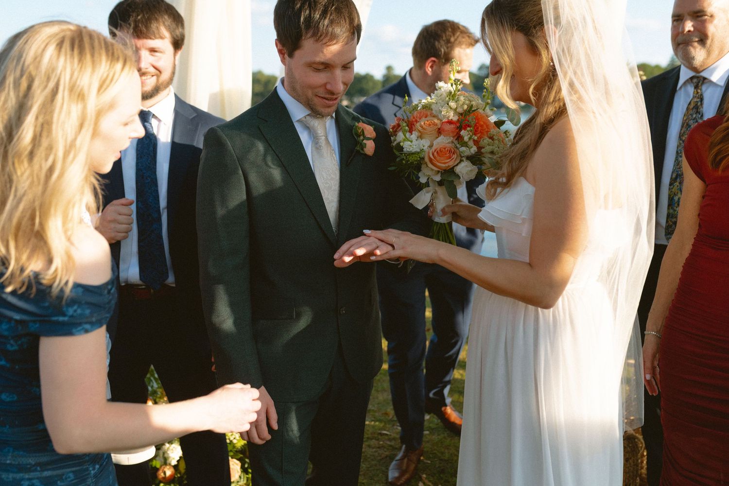 Groom examines wedding ring during outdoor ceremony while bride holds colorful floral bouquet.