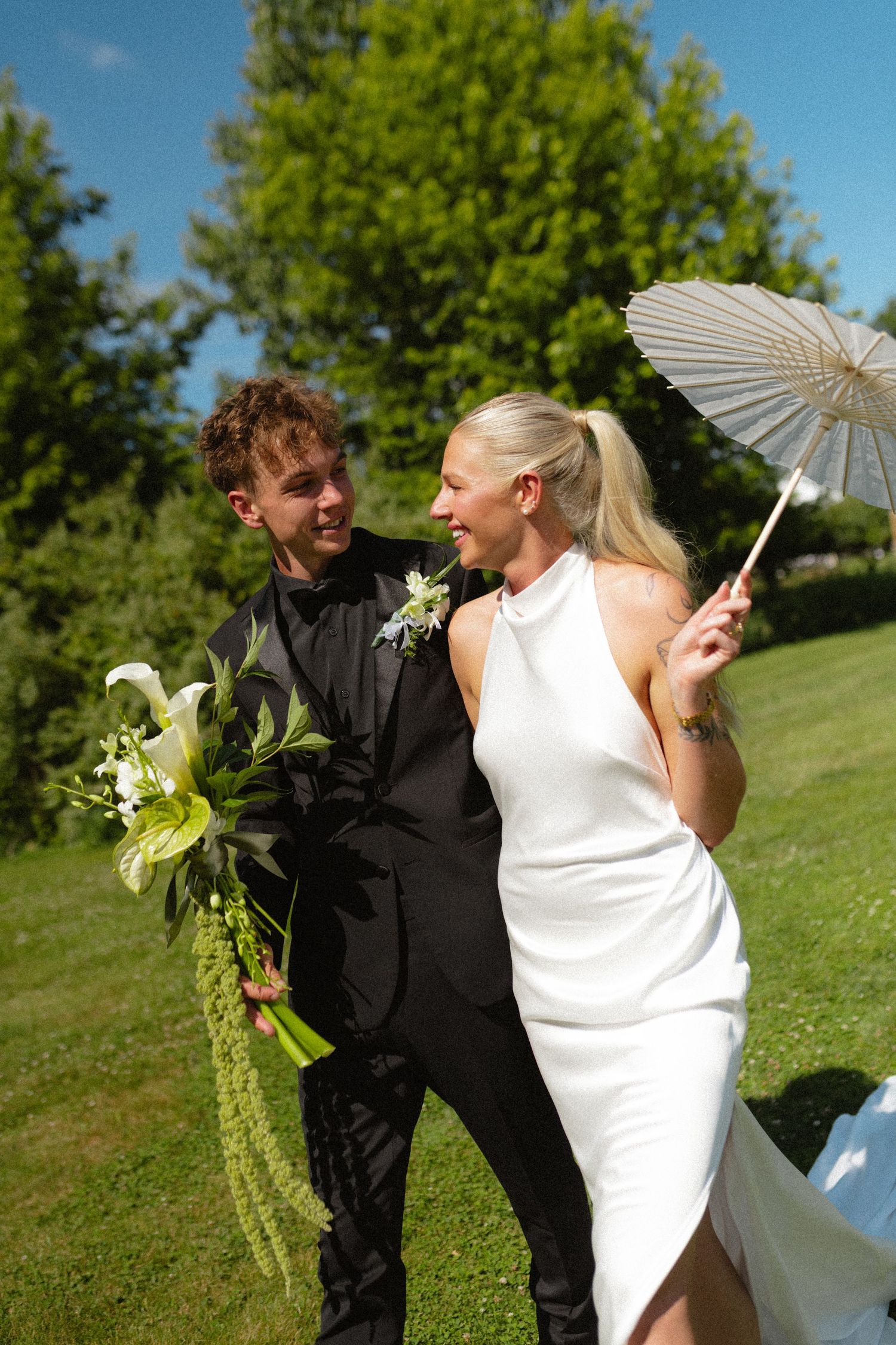 Bride in white dress holding parasol and groom in black suit walking in garden.