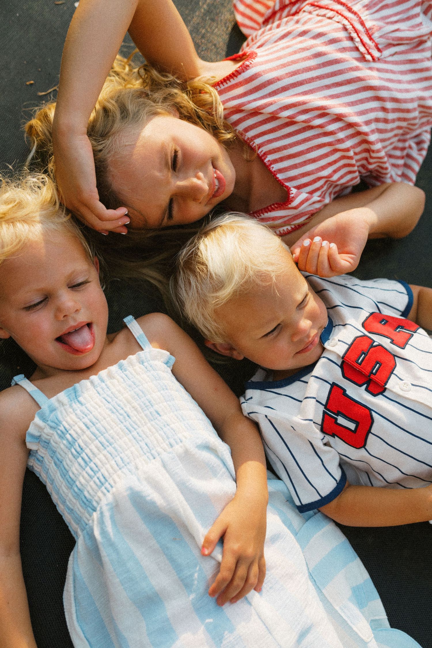 Three blonde siblings lying on pavement laughing and sticking out tongues on a sunny summer day.