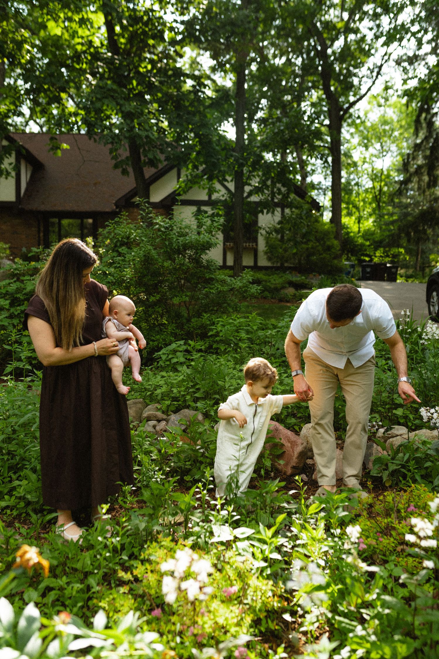 Family of four exploring a lush green garden, mother holding baby while father guides toddler among wildflowers.