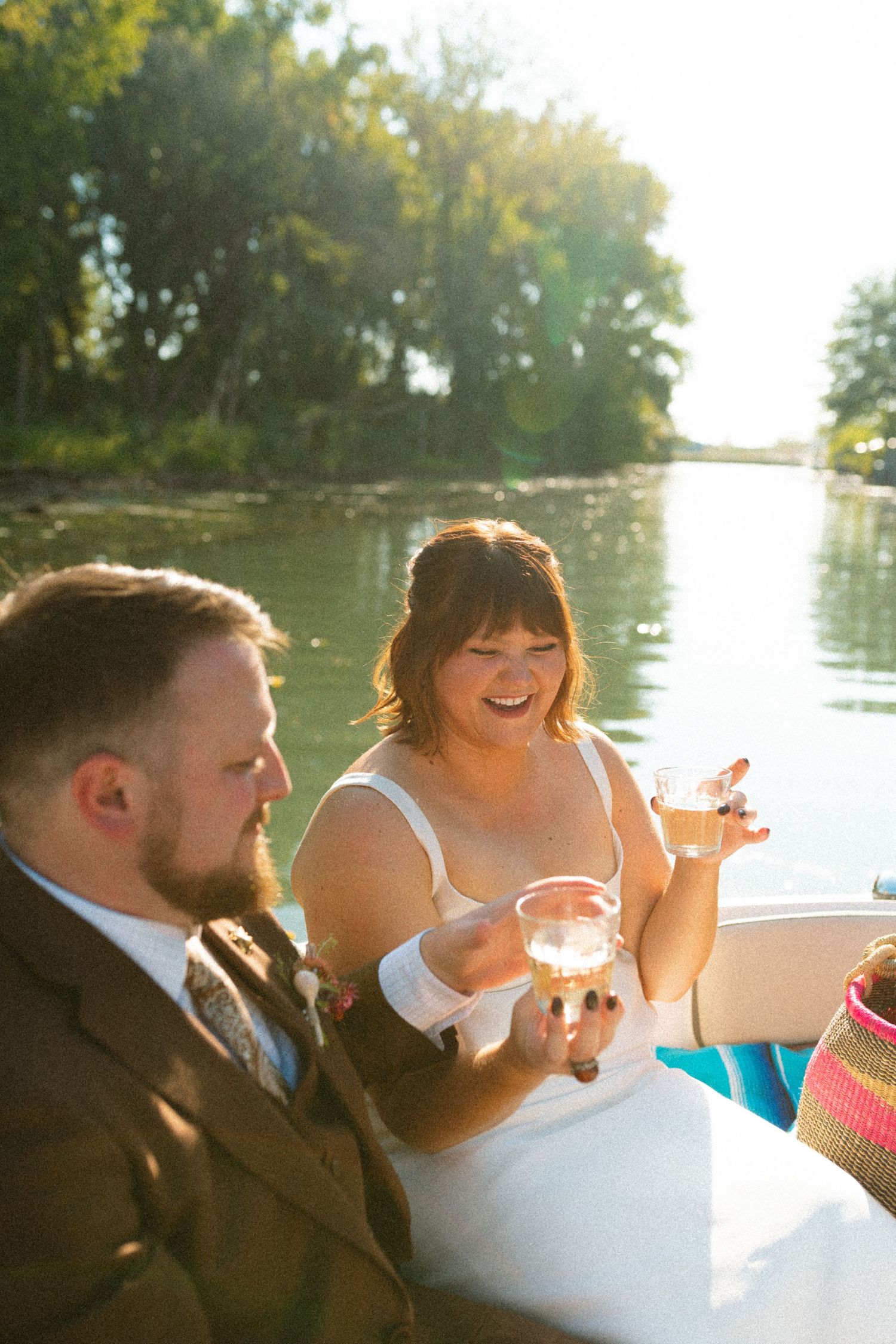 Bride and groom laughing and holding drinks while enjoying a romantic boat ride on a sunny lake.