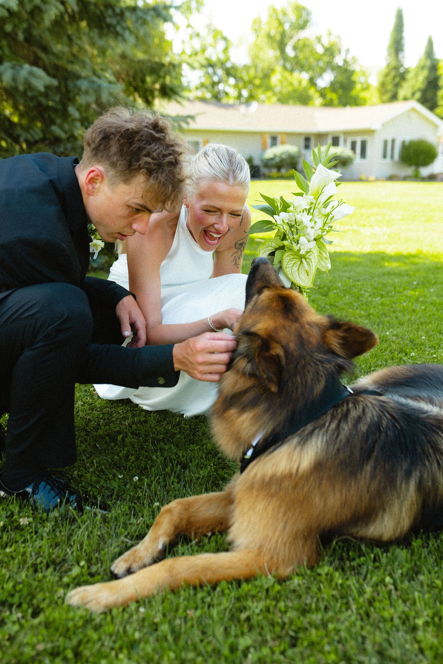 Bride and groom kneel on grass petting a German Shepherd dog.