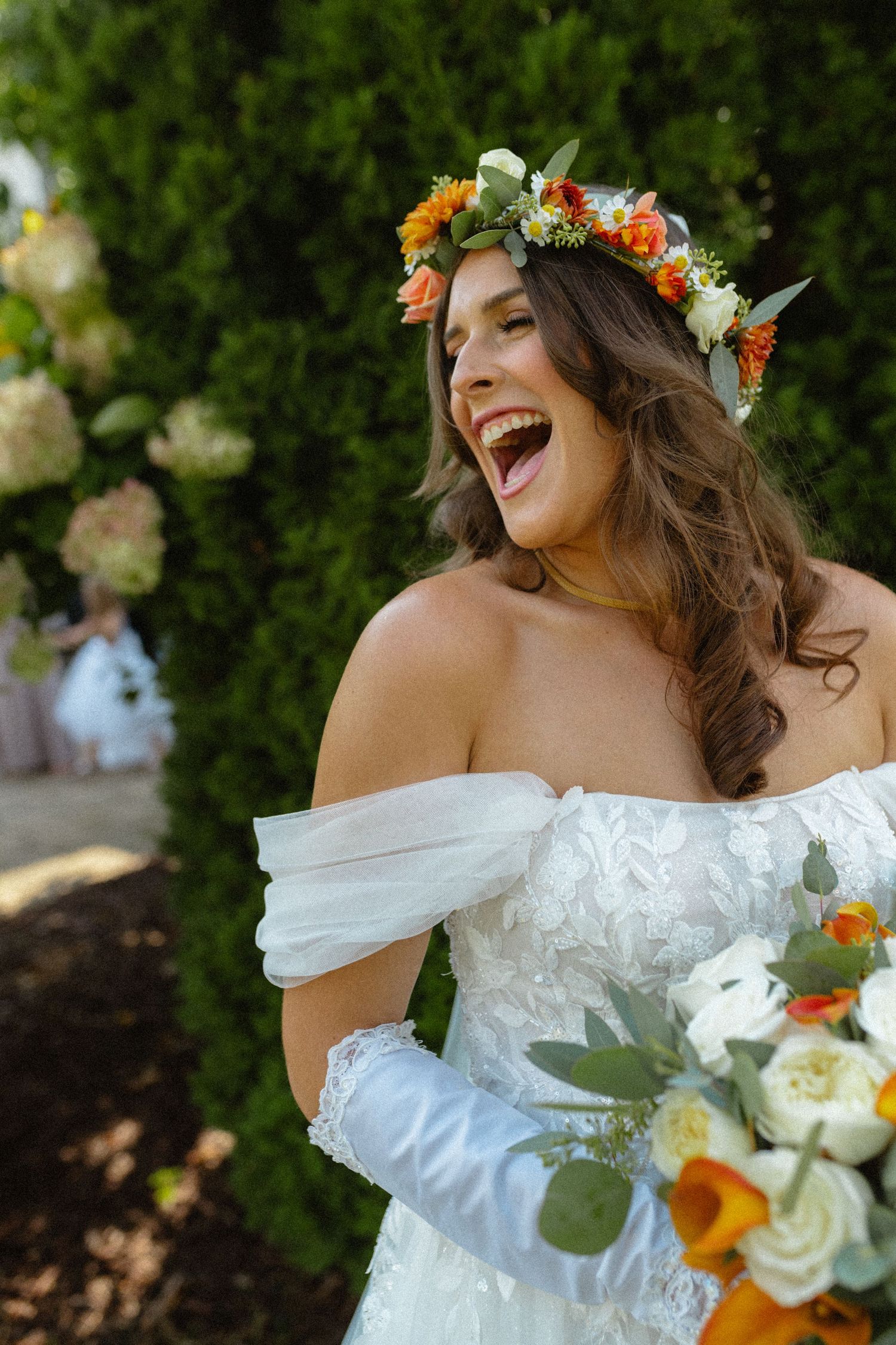 Joyful bride in floral crown and white off-shoulder dress holding colorful bouquet outdoors.