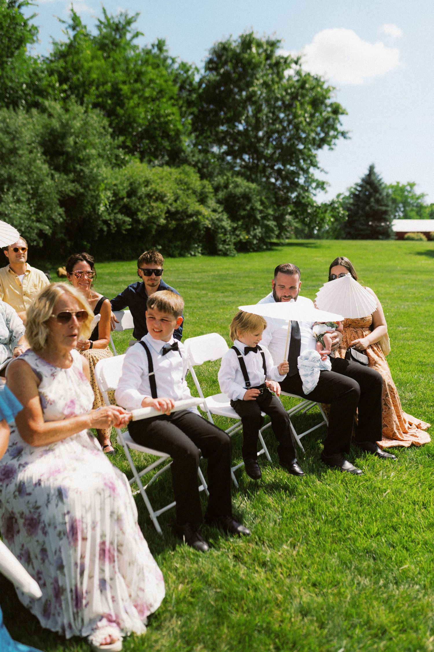 Guests seated on white chairs at an outdoor wedding ceremony on a sunny day with lush green lawn.