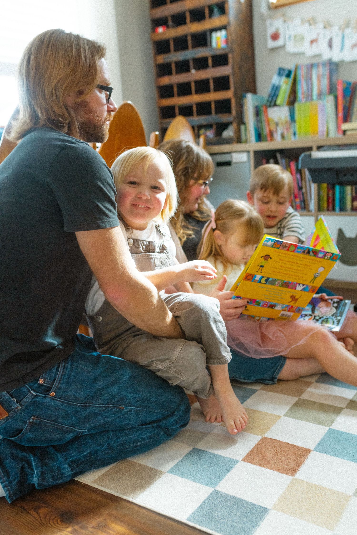 Father sitting on checkered rug reading a book with smiling toddler on his lap in a home library.