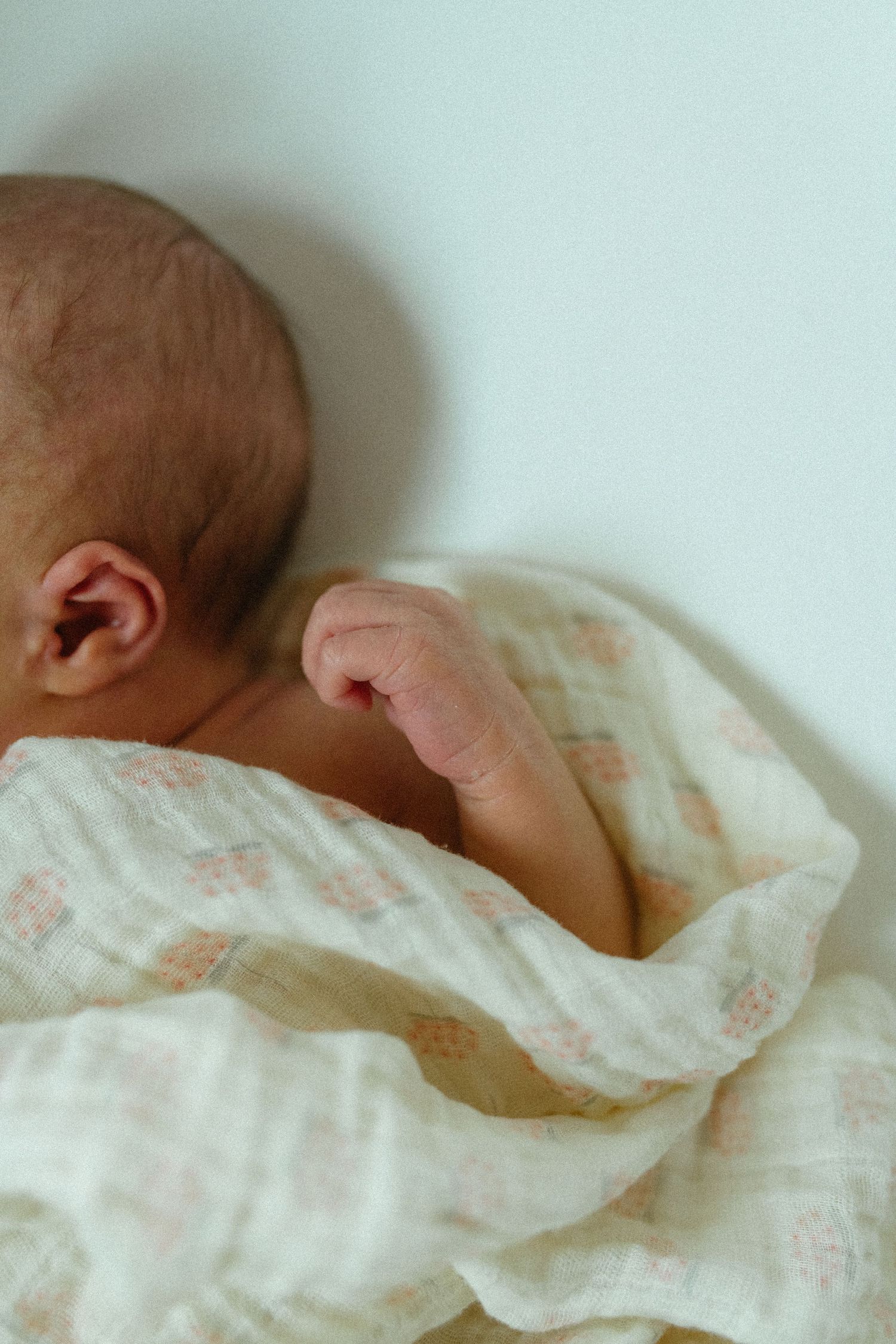 Sleeping newborn baby wrapped in floral muslin swaddle blanket on white surface.
