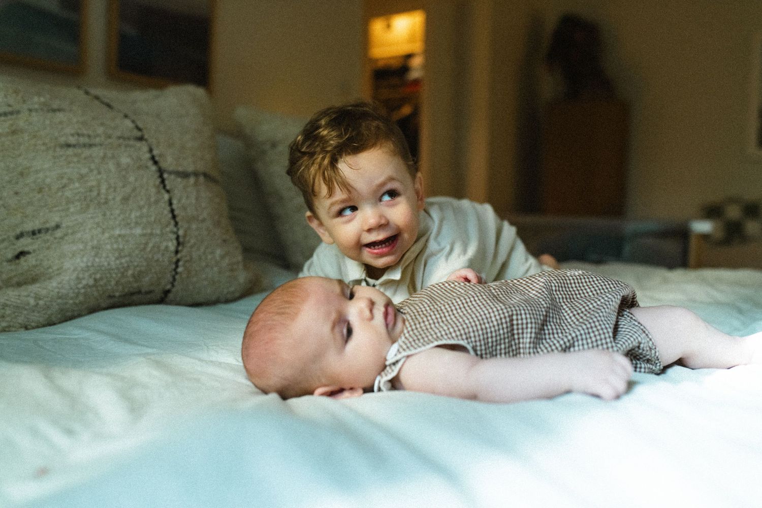 Happy toddler boy leaning over smiling baby sibling lying on a white bed indoors.