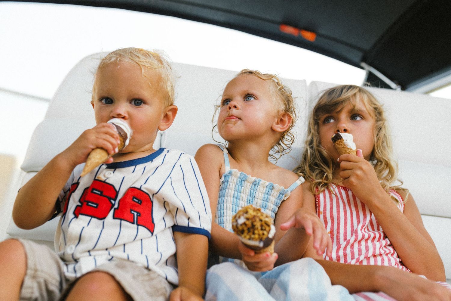Three young blonde children sitting together eating ice cream cones on a sunny summer day.