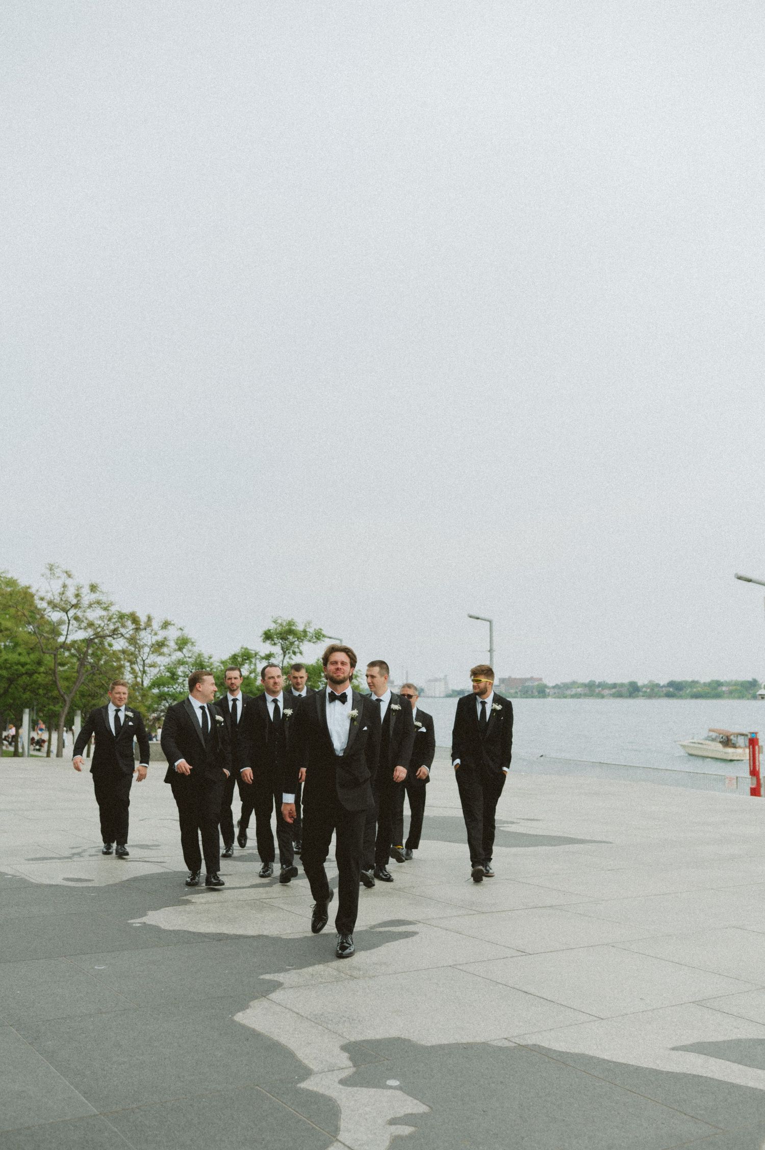Groom leads groomsmen in black tuxedos walking along waterfront plaza.