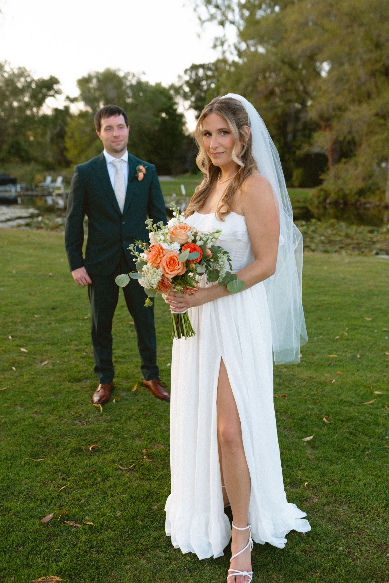 Bride in white gown with slit holds orange bouquet as groom in dark suit stands behind her.