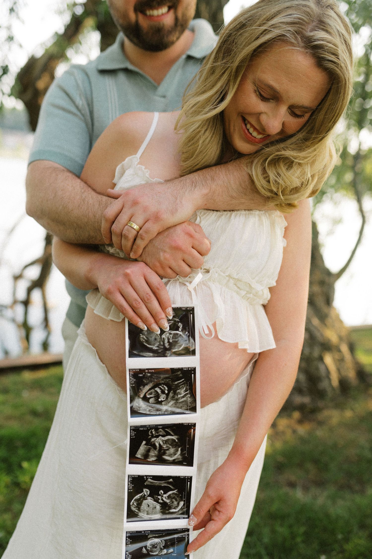 Smiling couple embracing outdoors holding ultrasound photos during maternity photo shoot.