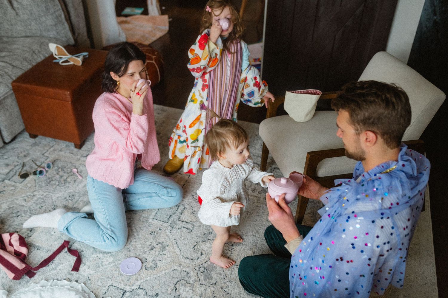Family of four sitting on living room floor playing with pink toy tea set together.