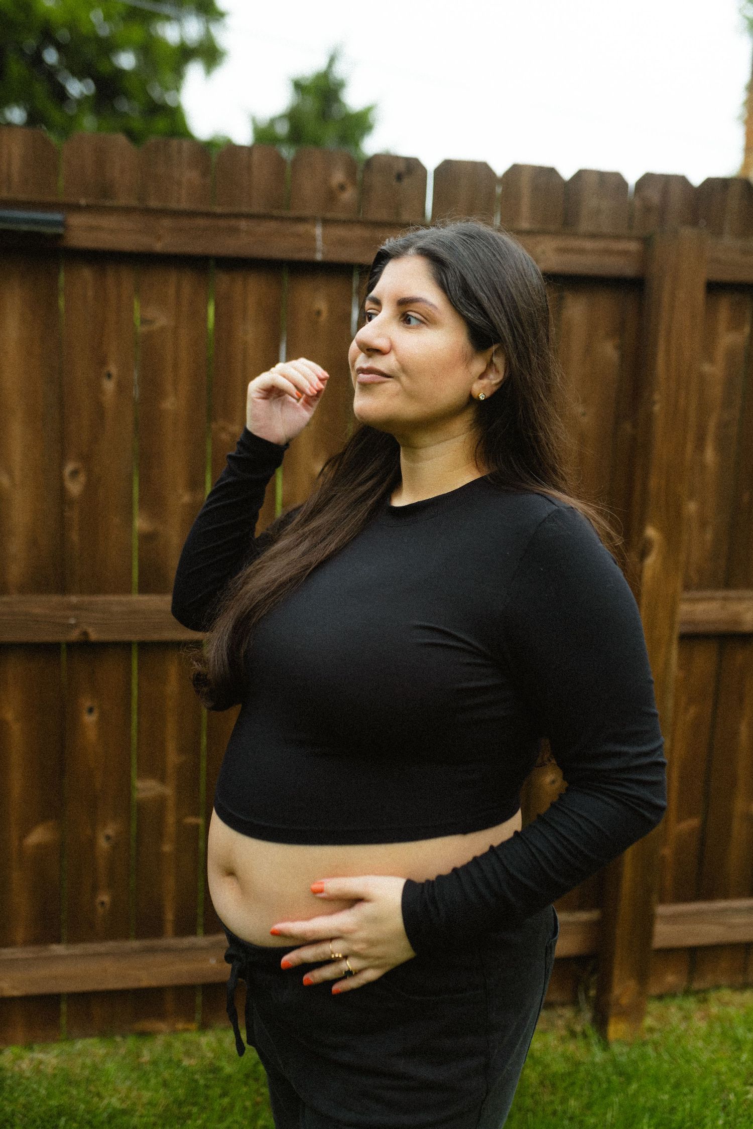 Pregnant woman in black crop top cradling baby bump outdoors near wooden fence.
