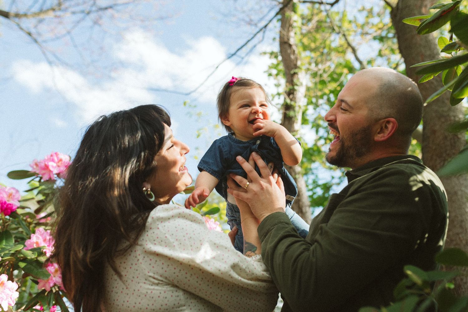 Joyful parents lifting and playing with their baby girl near blooming pink flowers outdoors.