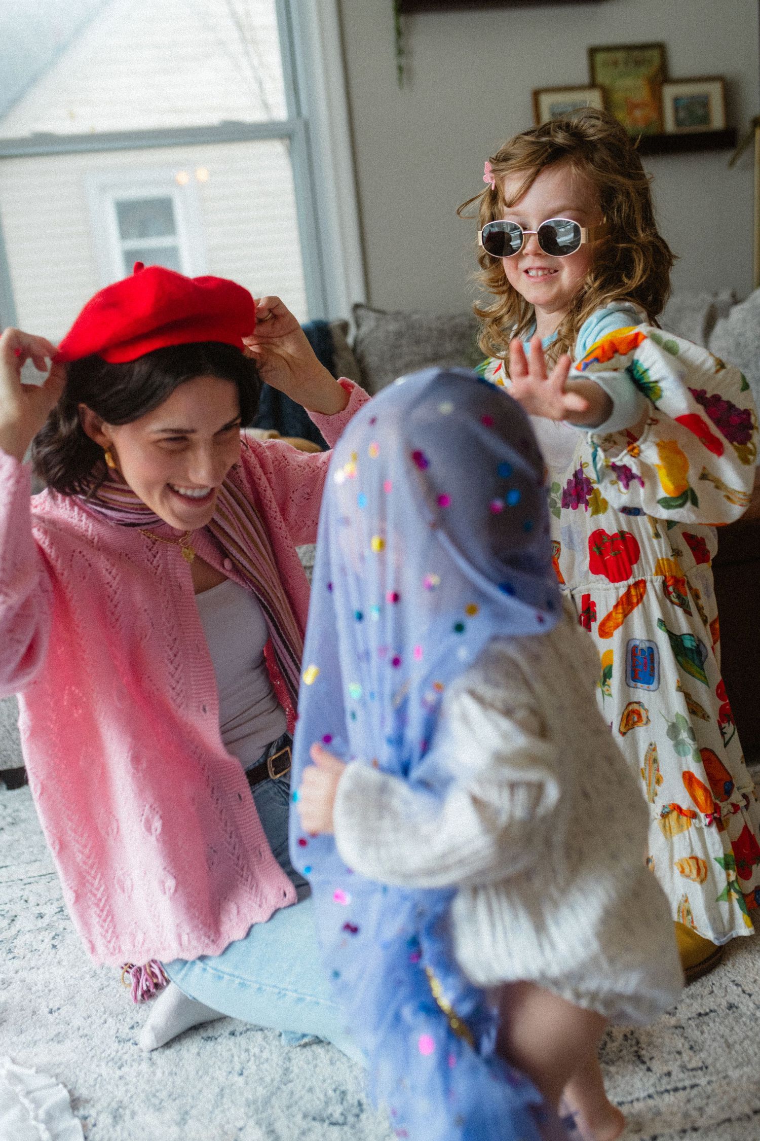 Woman in pink cardigan and red beret laughing with two young children in colorful outfits indoors.