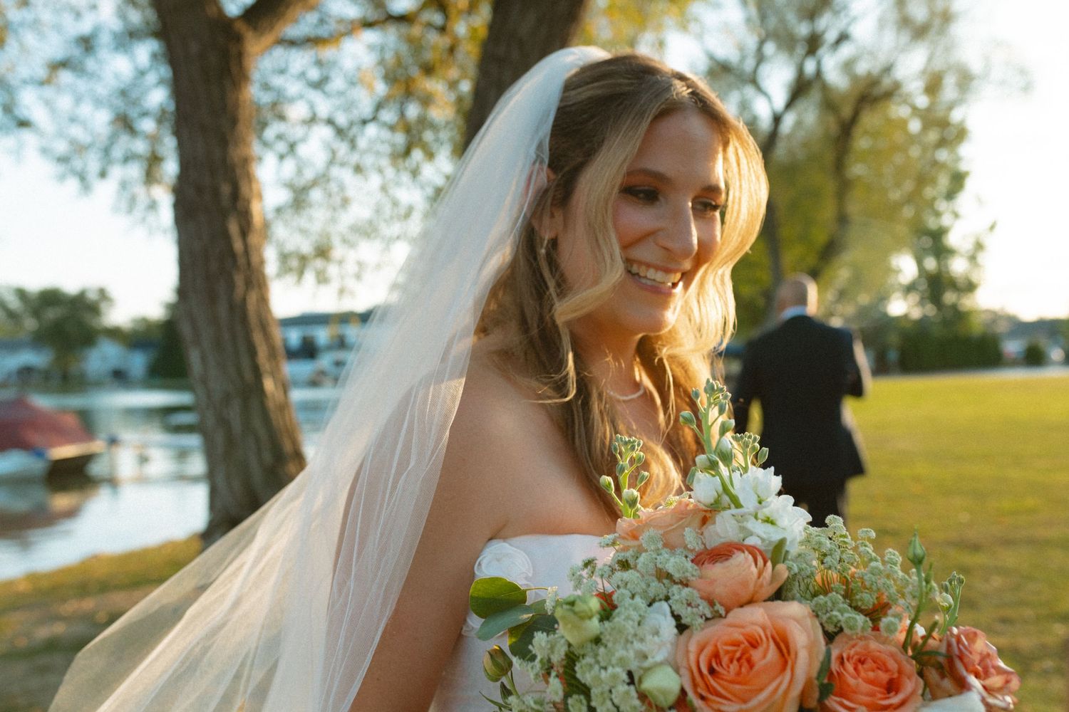 Smiling bride in white veil holds peach rose bouquet with sunlit lake and trees in background.