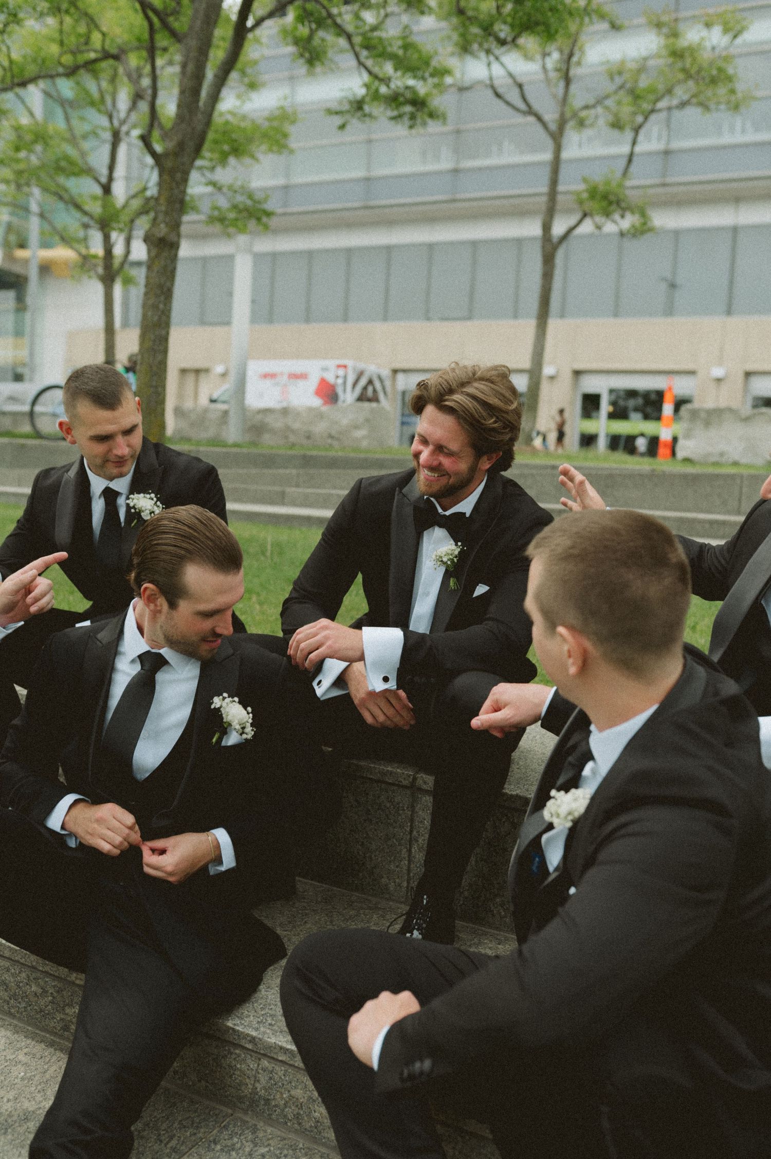 Groomsmen in black tuxedos with boutonnieres laugh together seated outdoors.