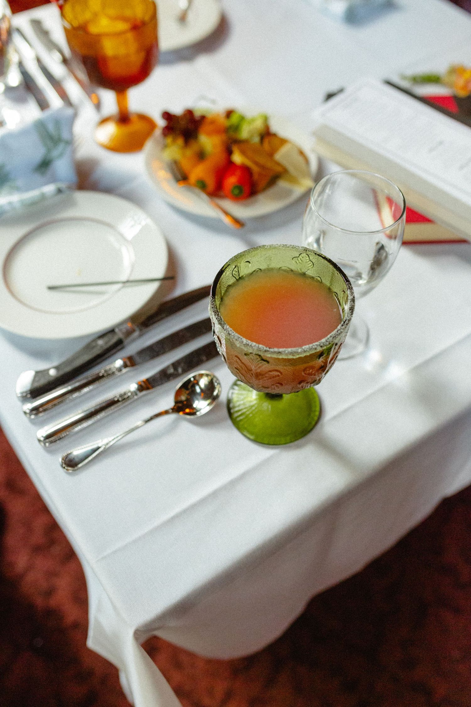 Elegant wedding reception table setting with green goblet, silverware, and colorful fruit appetizer plate.
