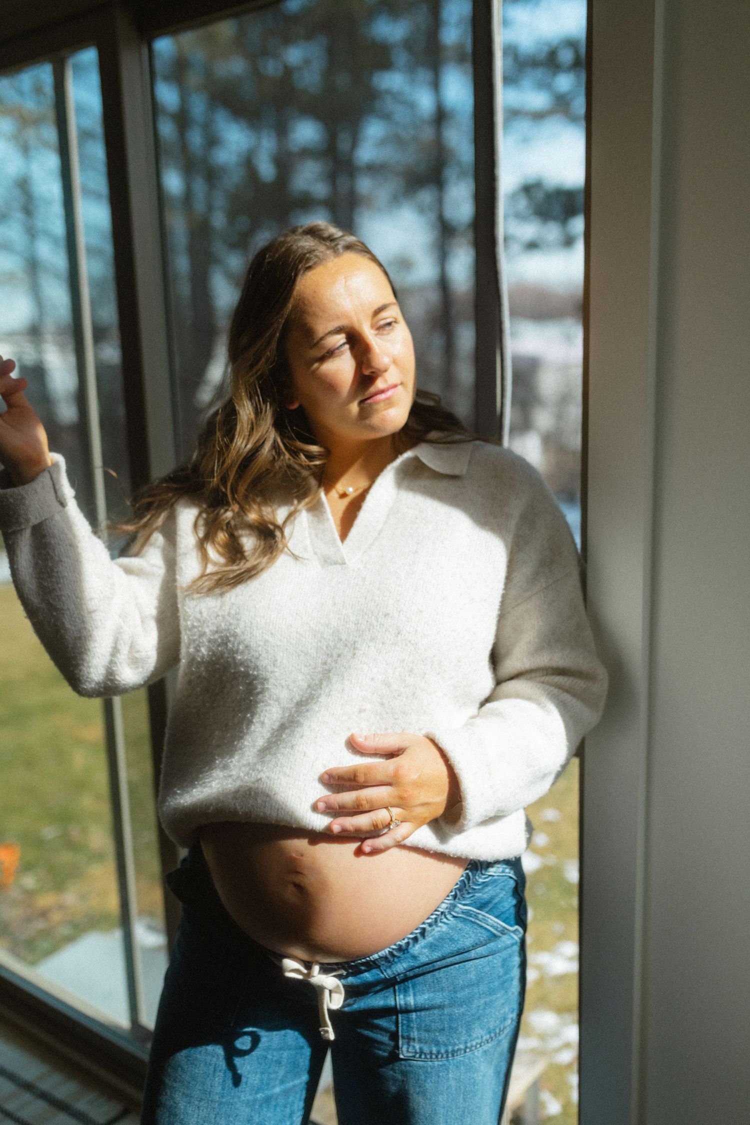 Pregnant woman in a white sweater and jeans holding her belly while standing by a sunny window.