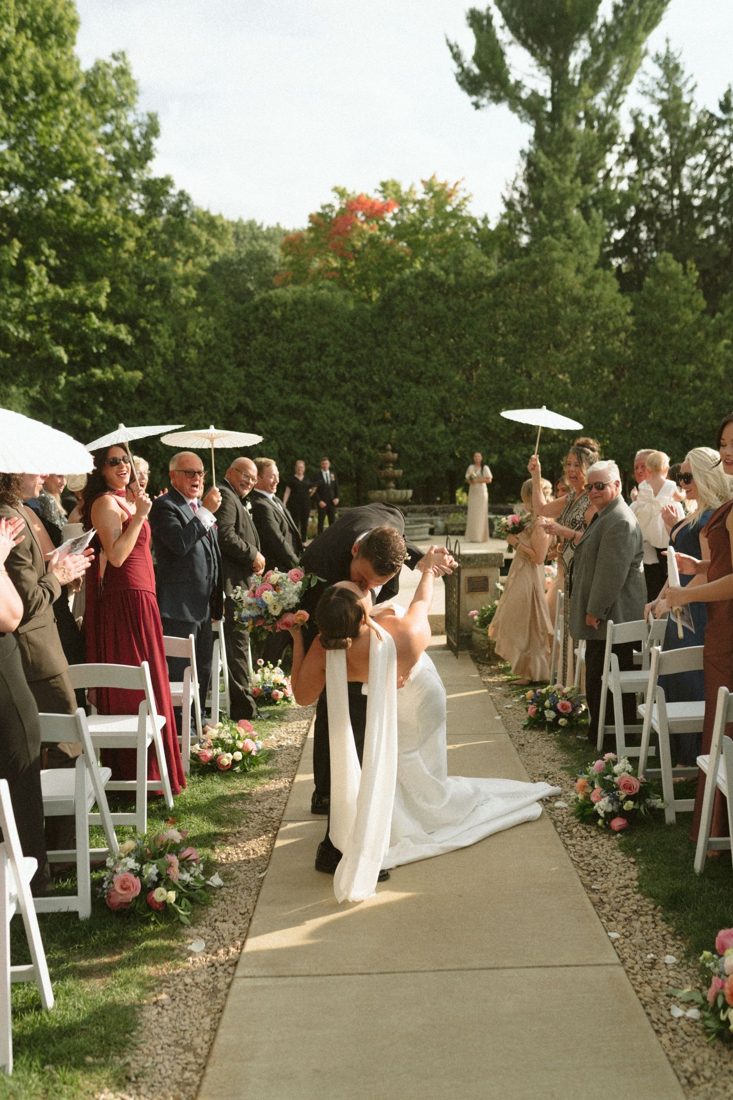 Bride and groom share romantic kiss in outdoor wedding ceremony aisle lined with white chairs.