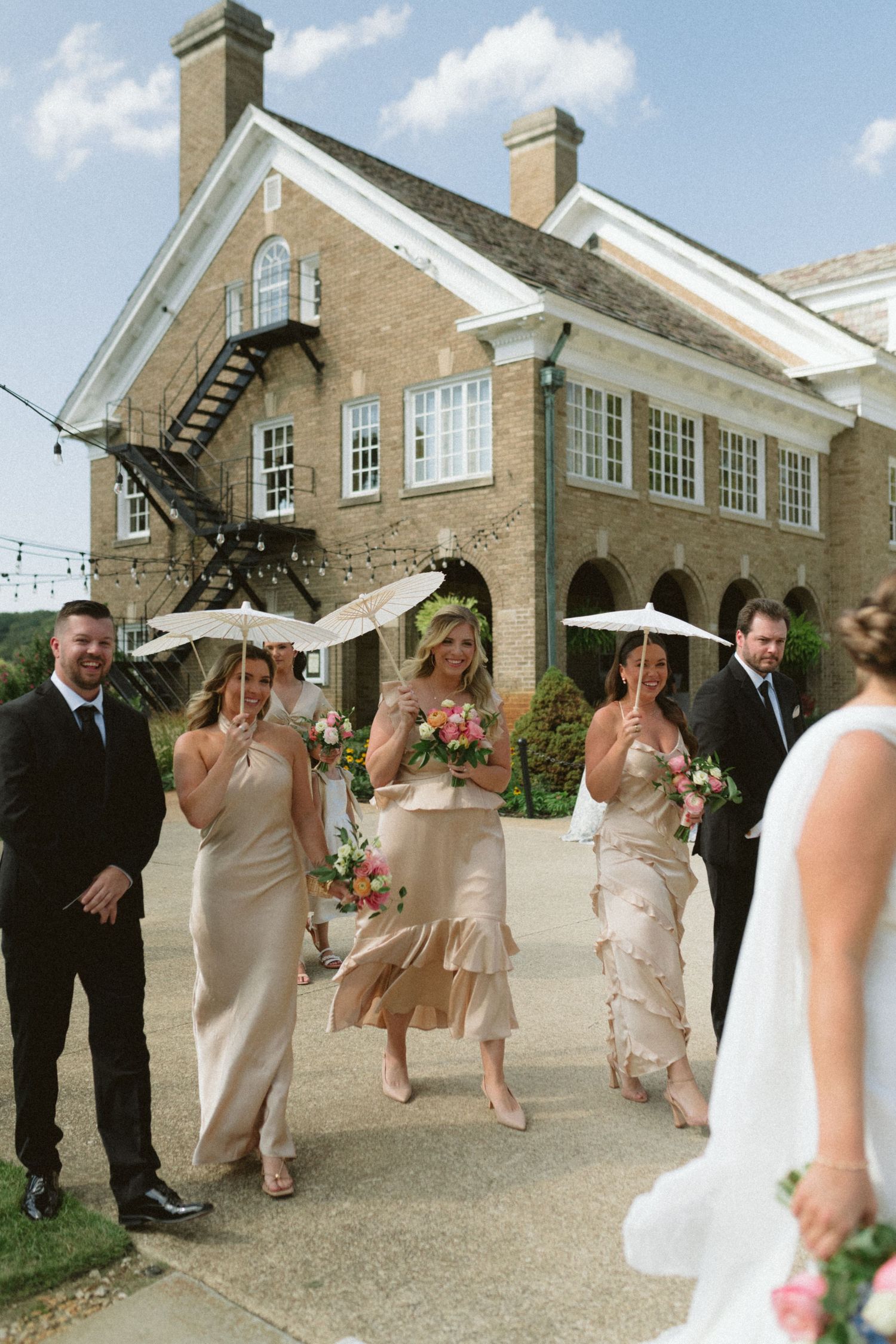 Bridesmaids in champagne gowns carrying parasols and bouquets walk toward bride at venue.