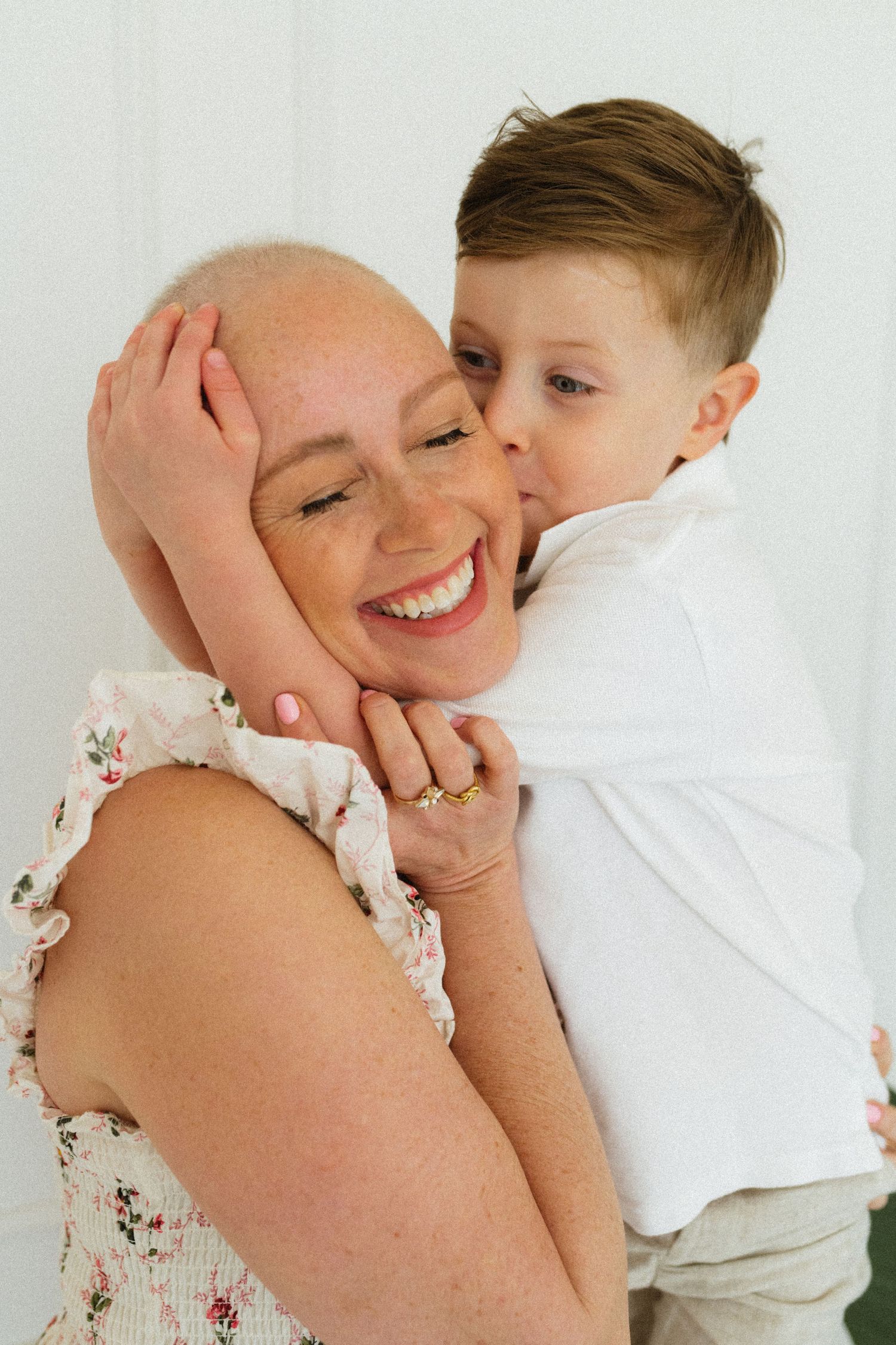 Young boy hugging and kissing a smiling bald woman in a floral dress.
