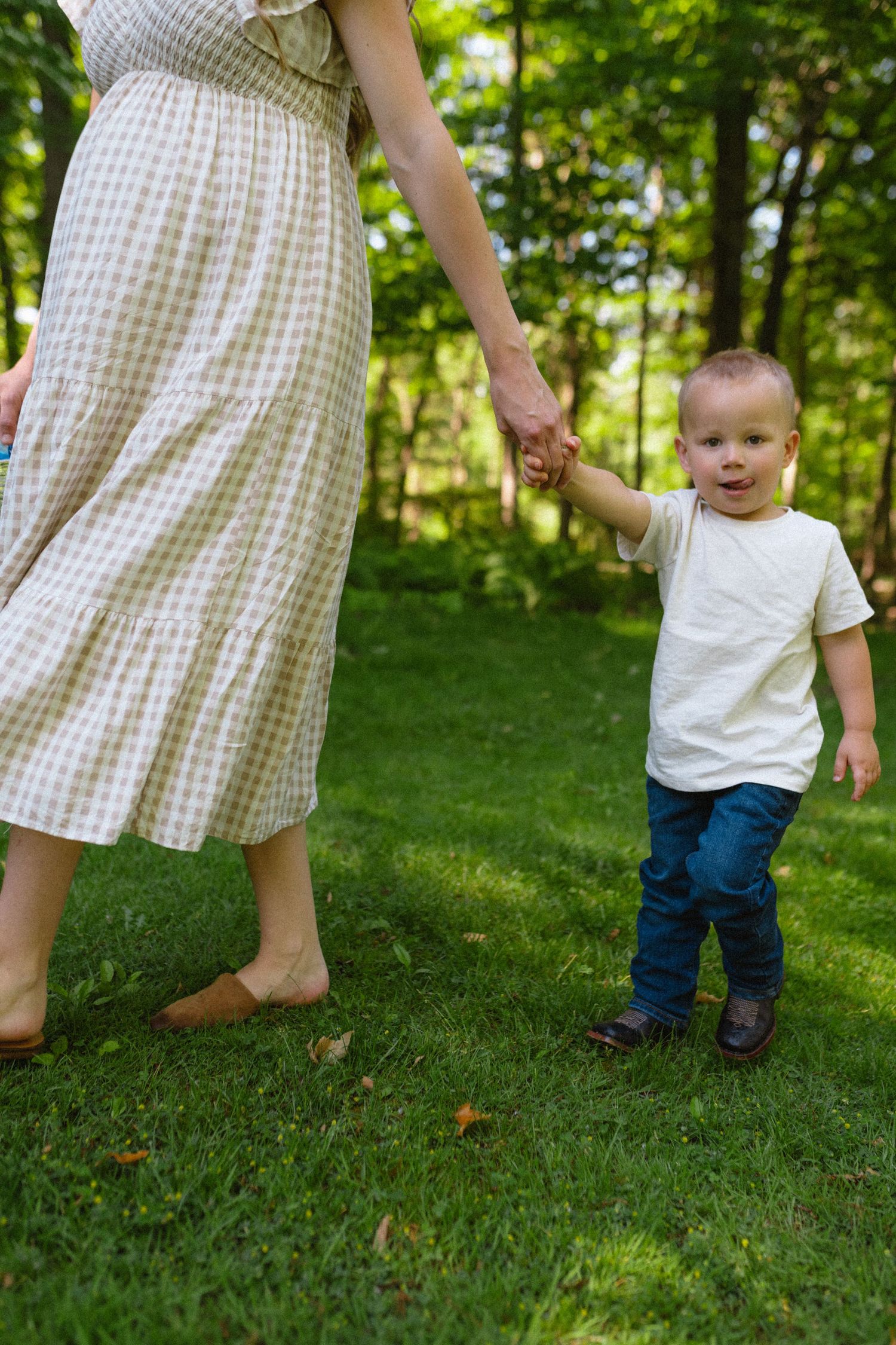 Toddler boy in white shirt holding mothers hand walking on green grass in wooded park.