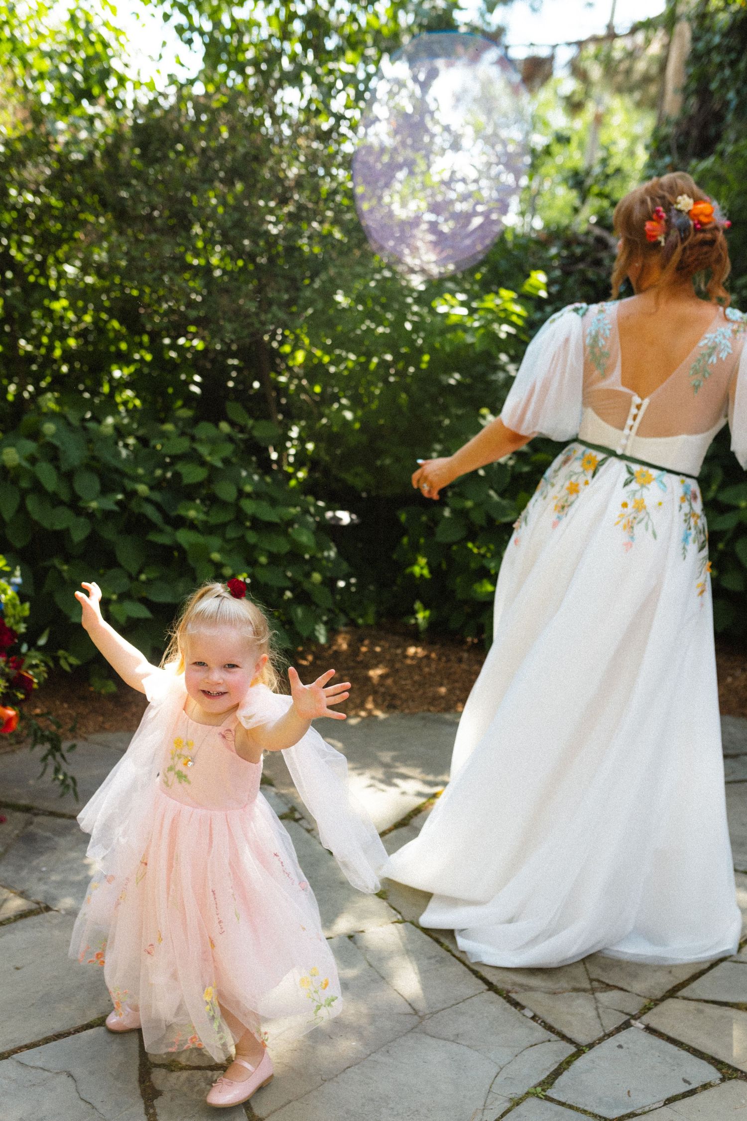 Joyful flower girl in pink dress with arms raised beside bride in floral wedding gown in garden.