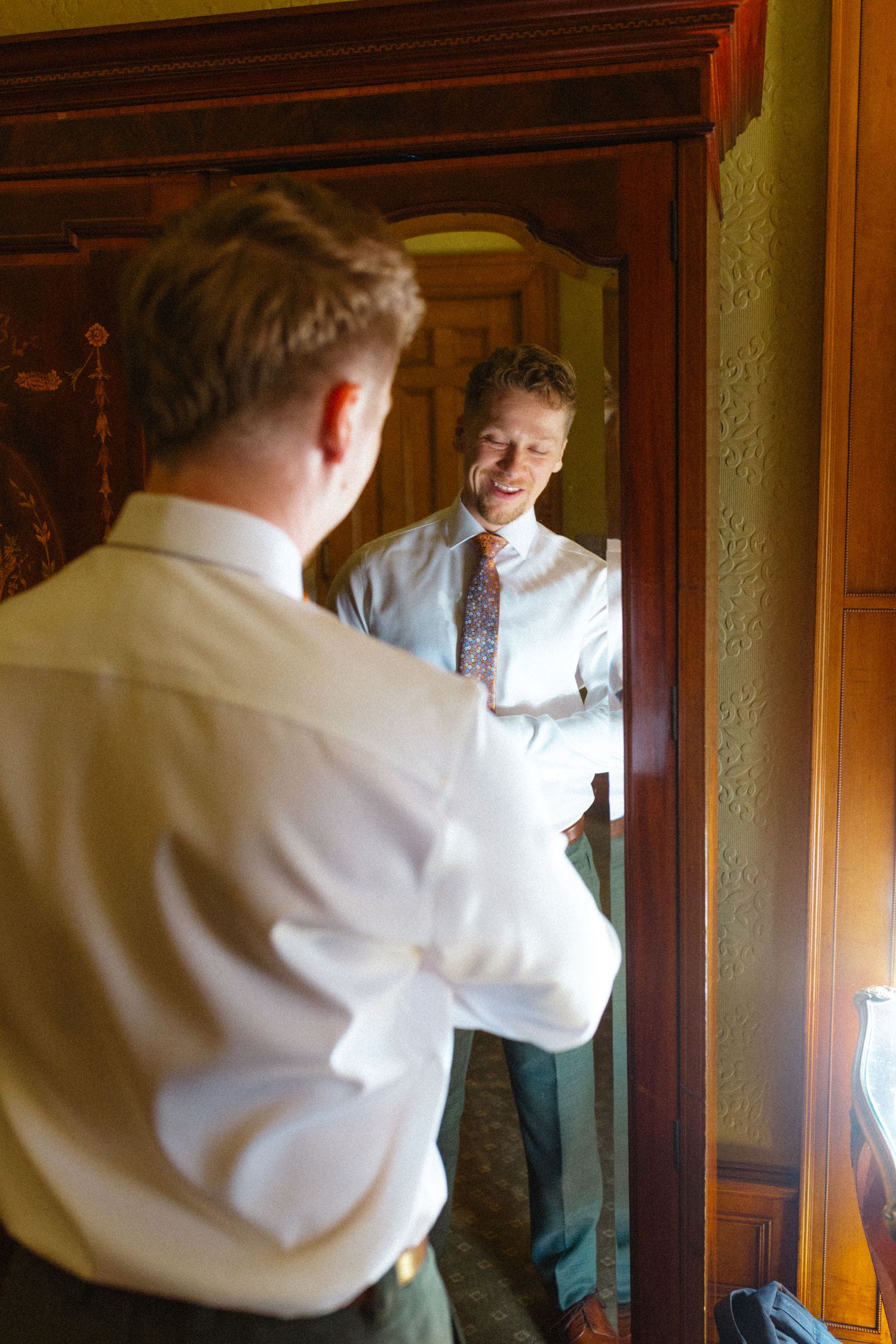 Groom smiles at his reflection while dressing in ornate mirror.