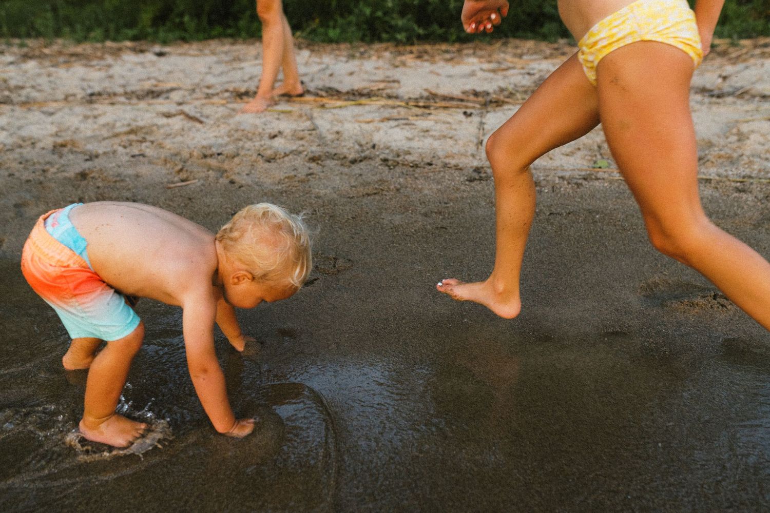 Toddler in colorful swim trunks bending down to touch wet sand while a child runs nearby.