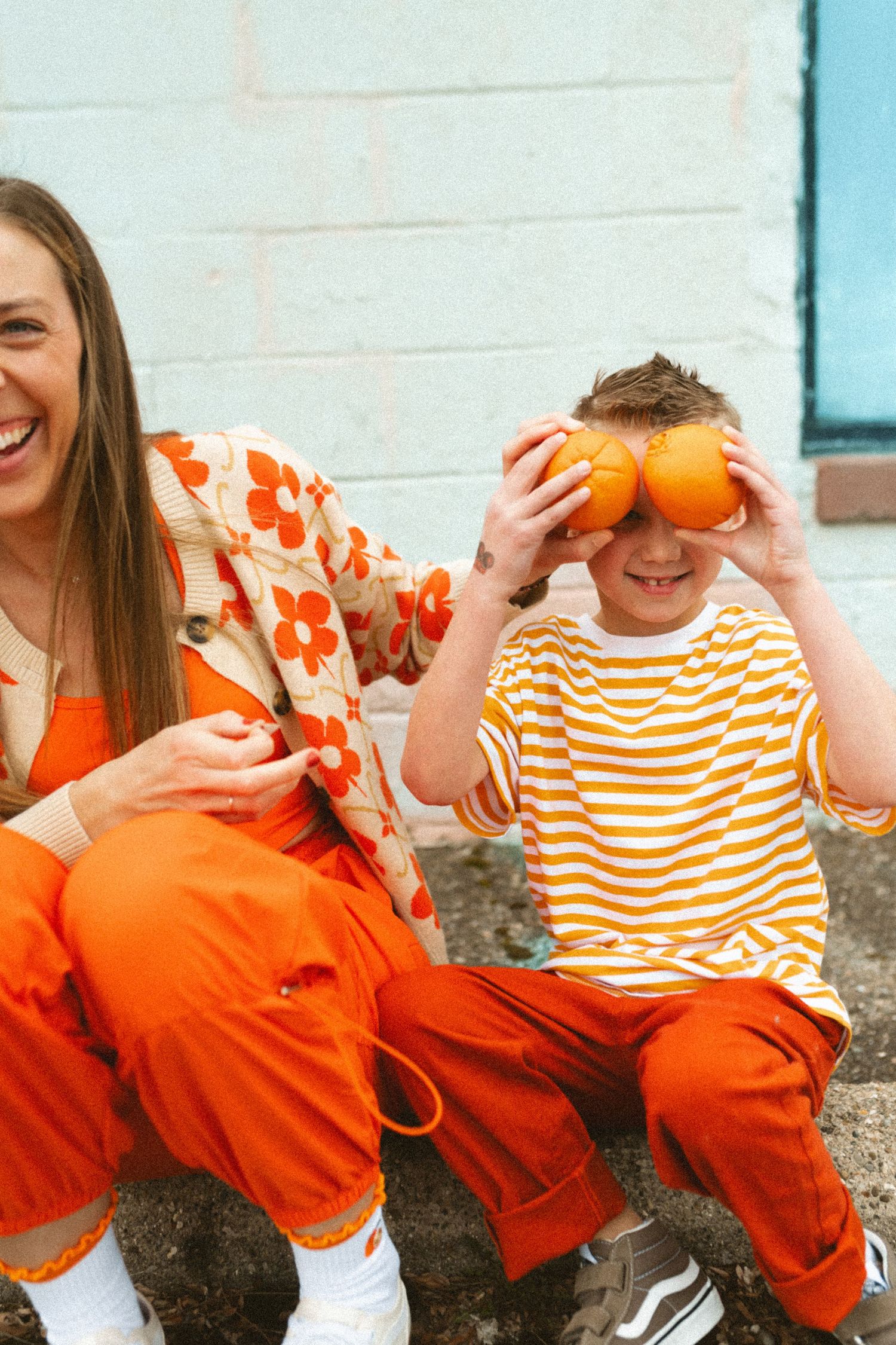 Laughing woman in orange floral cardigan beside boy holding oranges over his eyes.