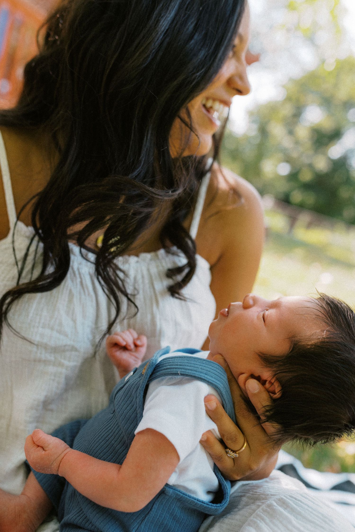Smiling mother with dark hair gazing lovingly at sleeping newborn baby outdoors in natural light.