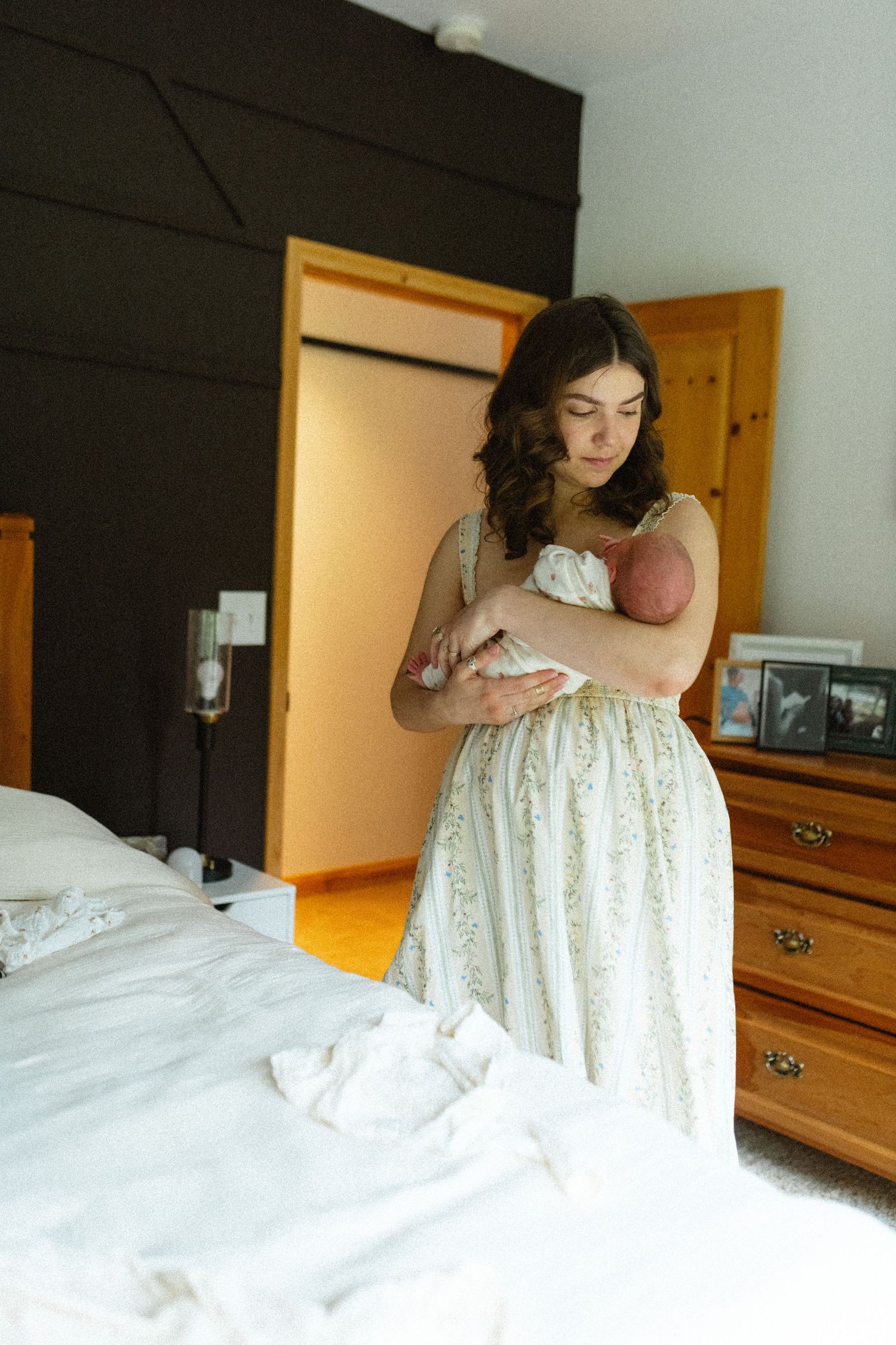 A mother in a white floral dress tenderly holds her newborn baby in a warmly decorated bedroom.
